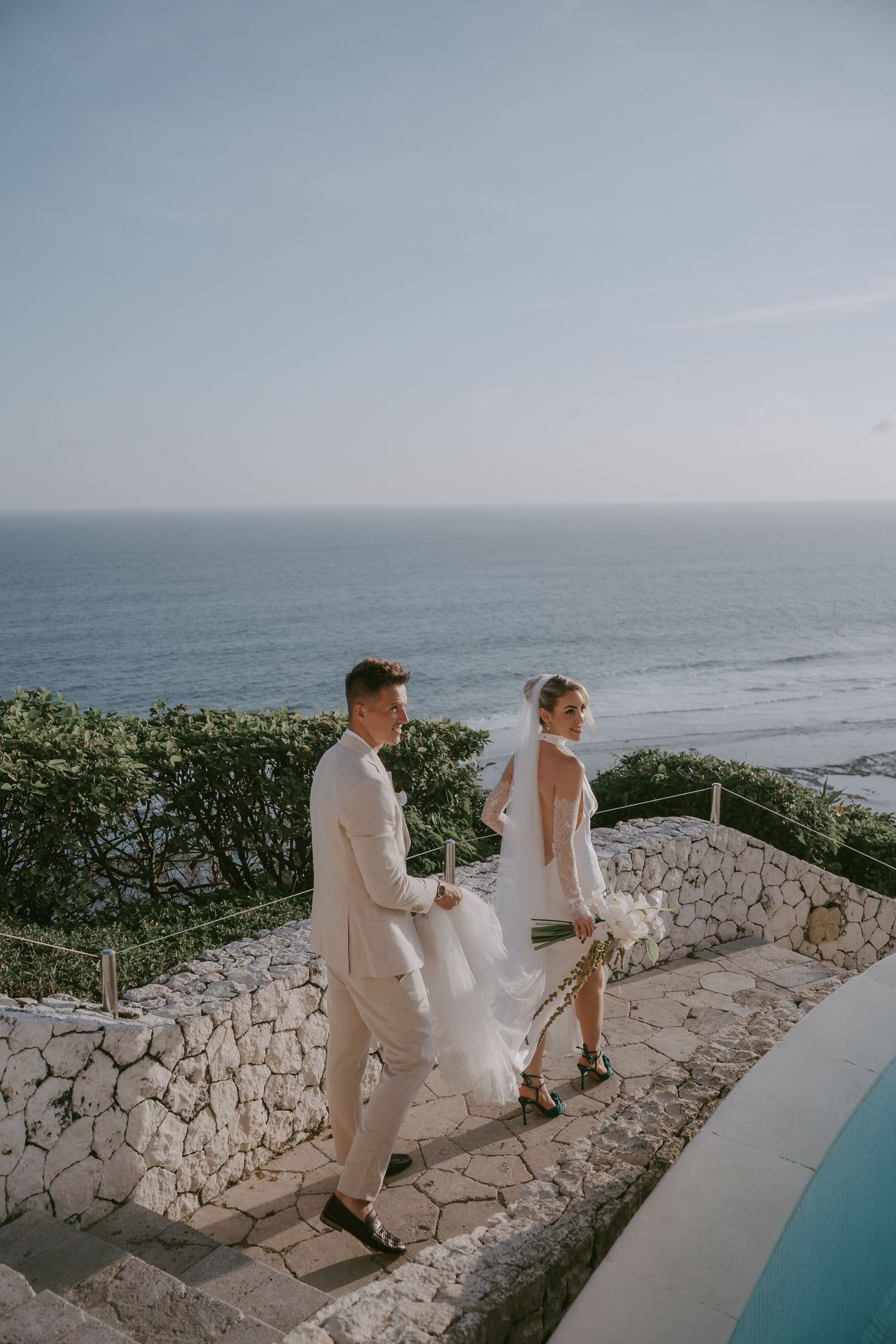 Couple walks hand in hand along stone pathway overlooking ocean and infinity pool.