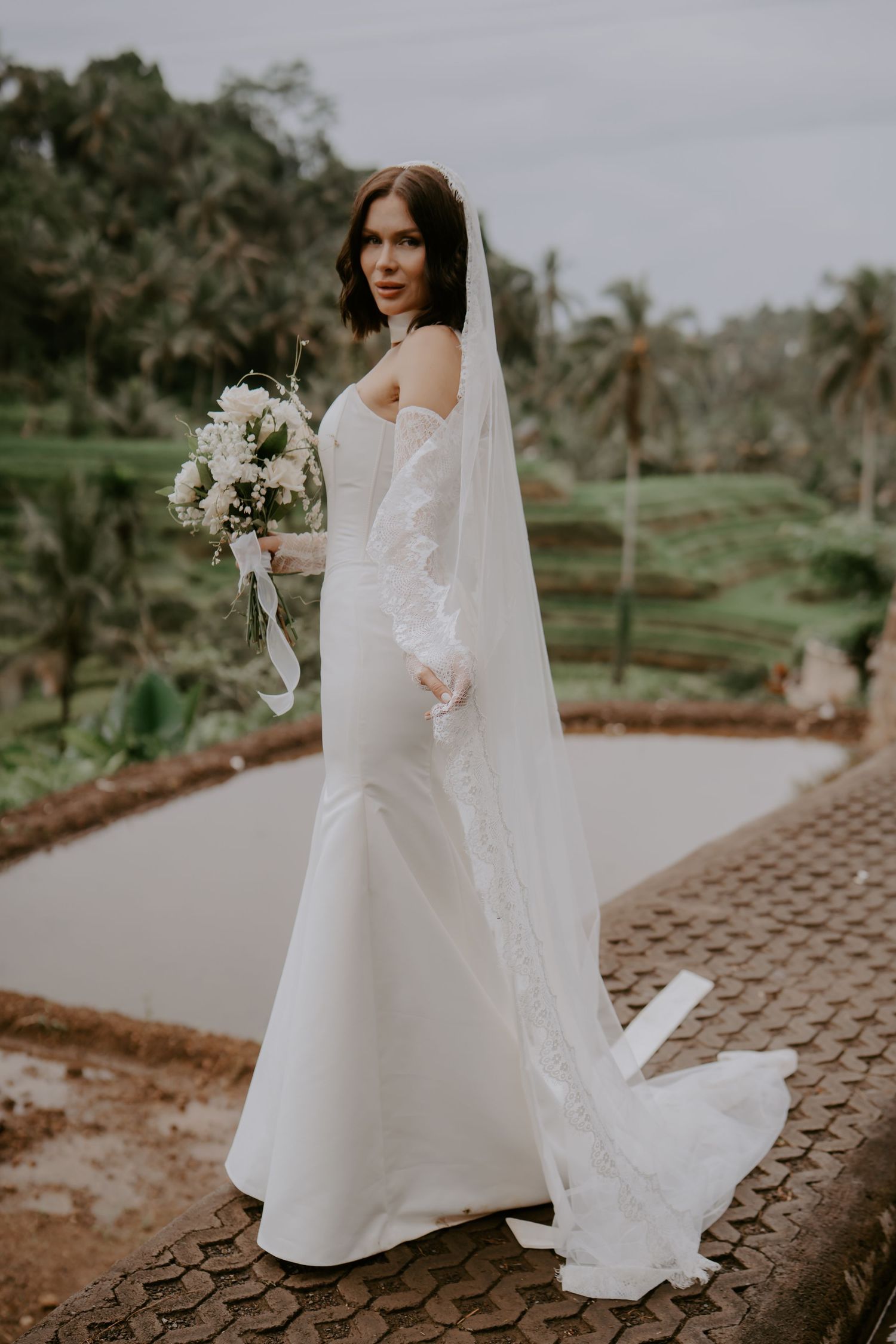 A bride in an elegant white off-shoulder wedding gown with long veil holds a bouquet against a tropical landscape backdrop.