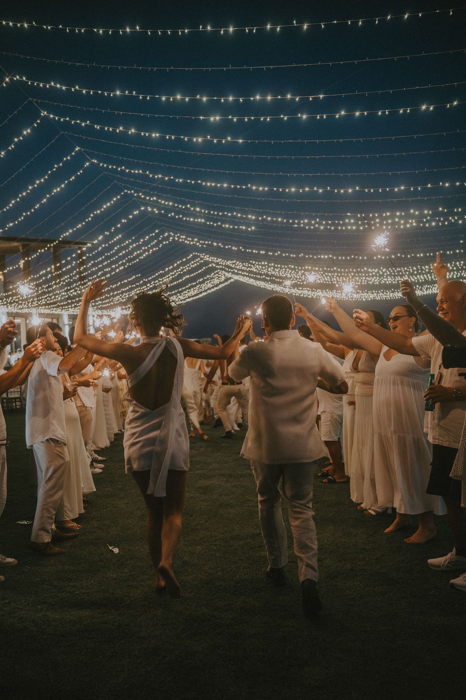 People dancing and celebrating at an outdoor event under strings of twinkling lights at night.