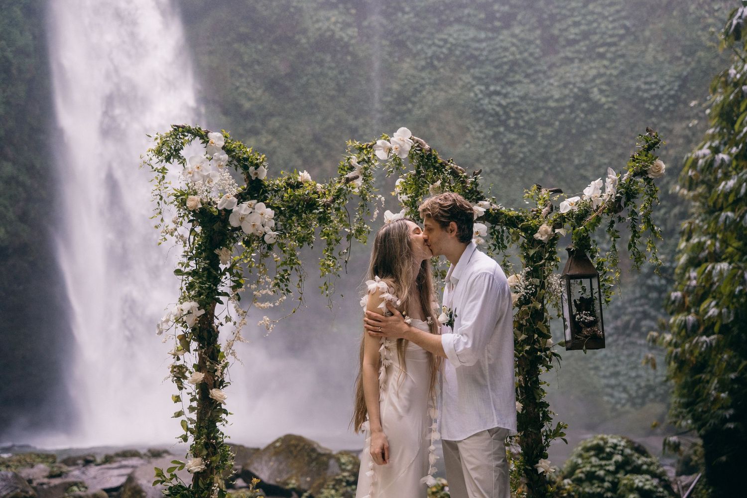 Couple embraces near a majestic waterfall surrounded by lush greenery and mist.