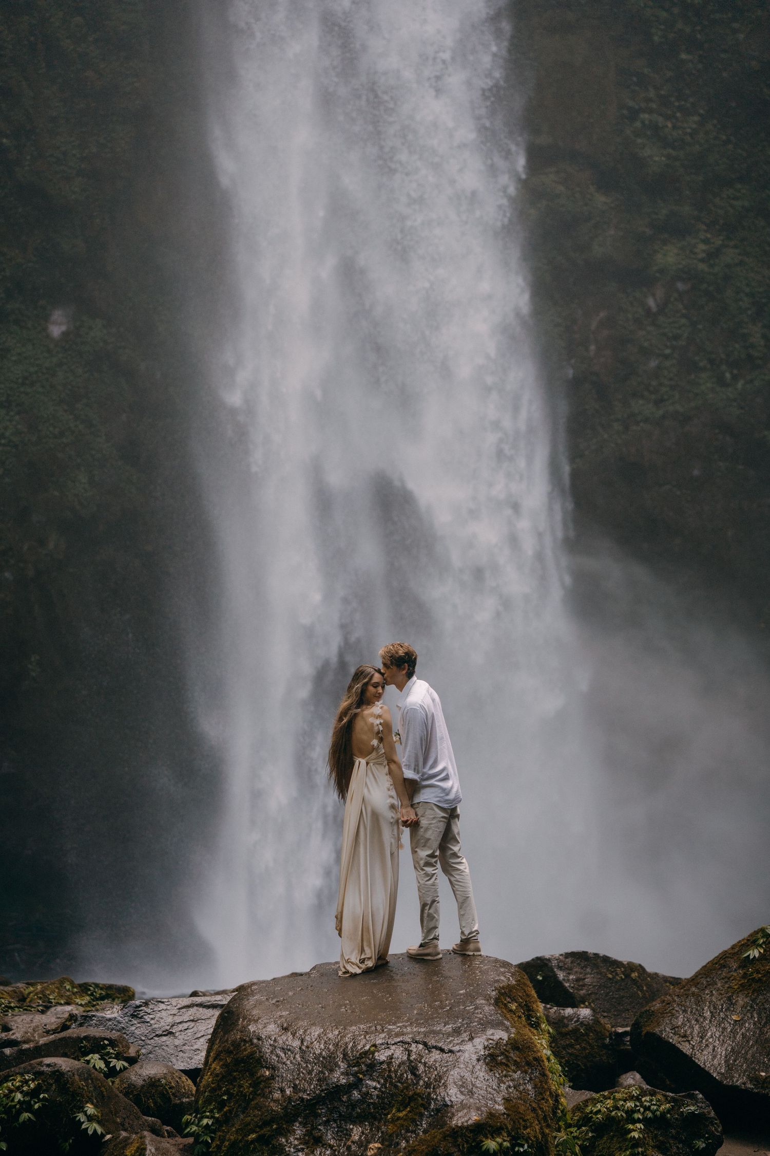 A couple stands together on a rocky outcrop near a majestic cascading waterfall in a misty natural setting.