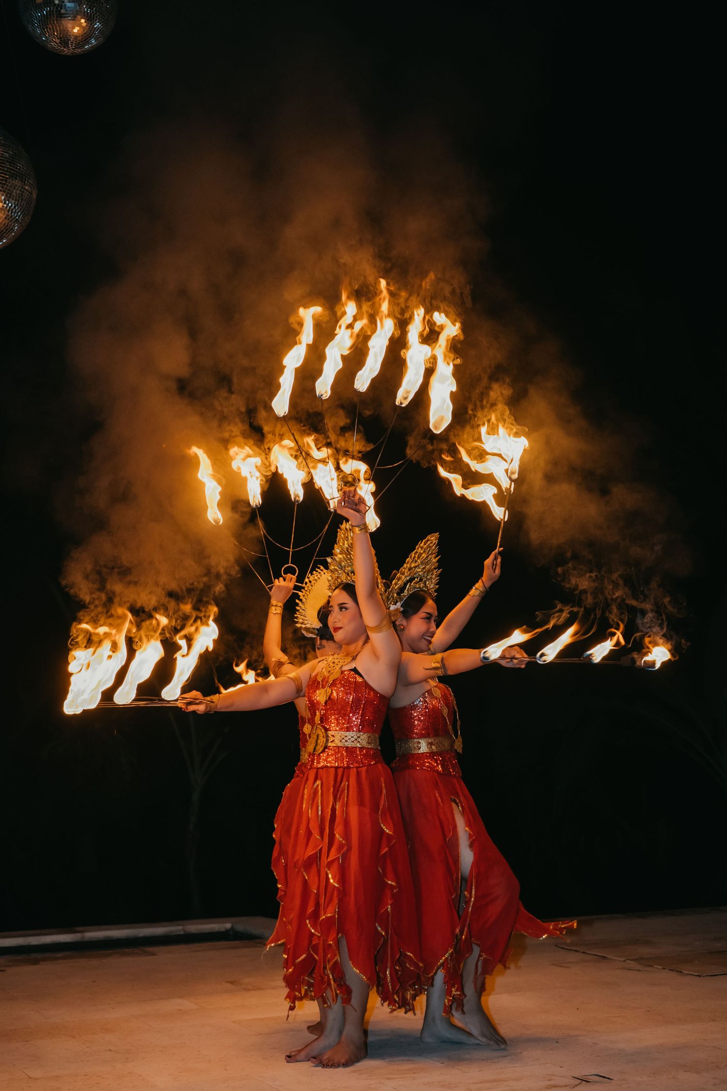 Two female fire dancers in red costumes performing with flaming props against a dramatic smoke-filled dark background.