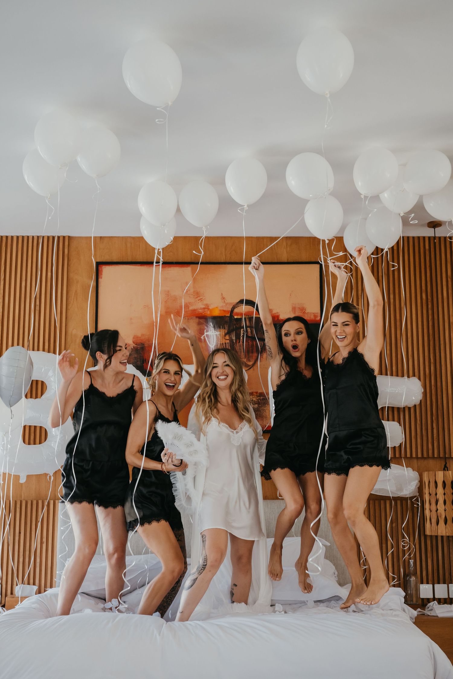 Bride and bridesmaids in black pajamas jumping on bed with white balloons at bachelorette party.