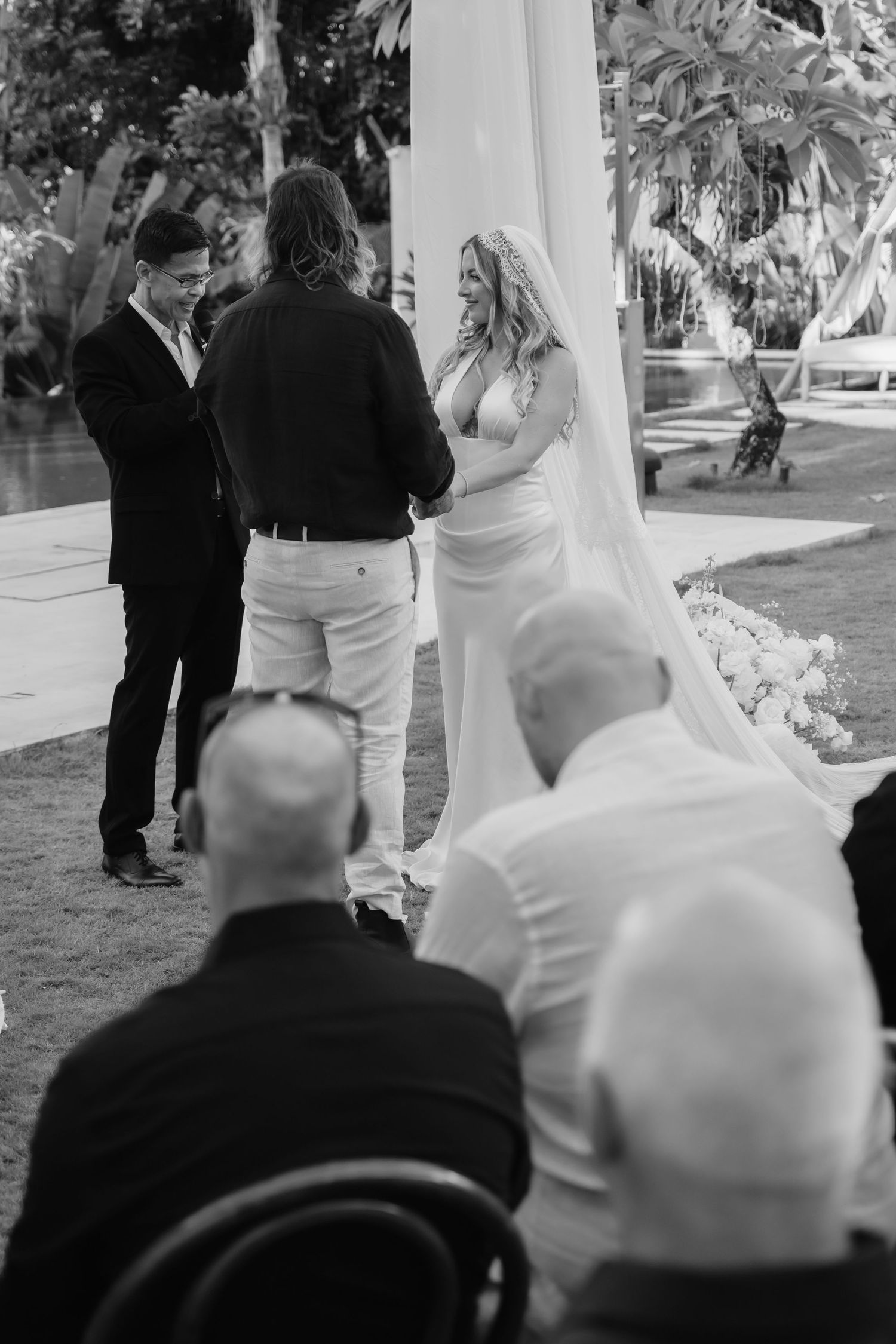 Black and white photo of bride and groom exchanging vows with officiant before seated wedding guests.