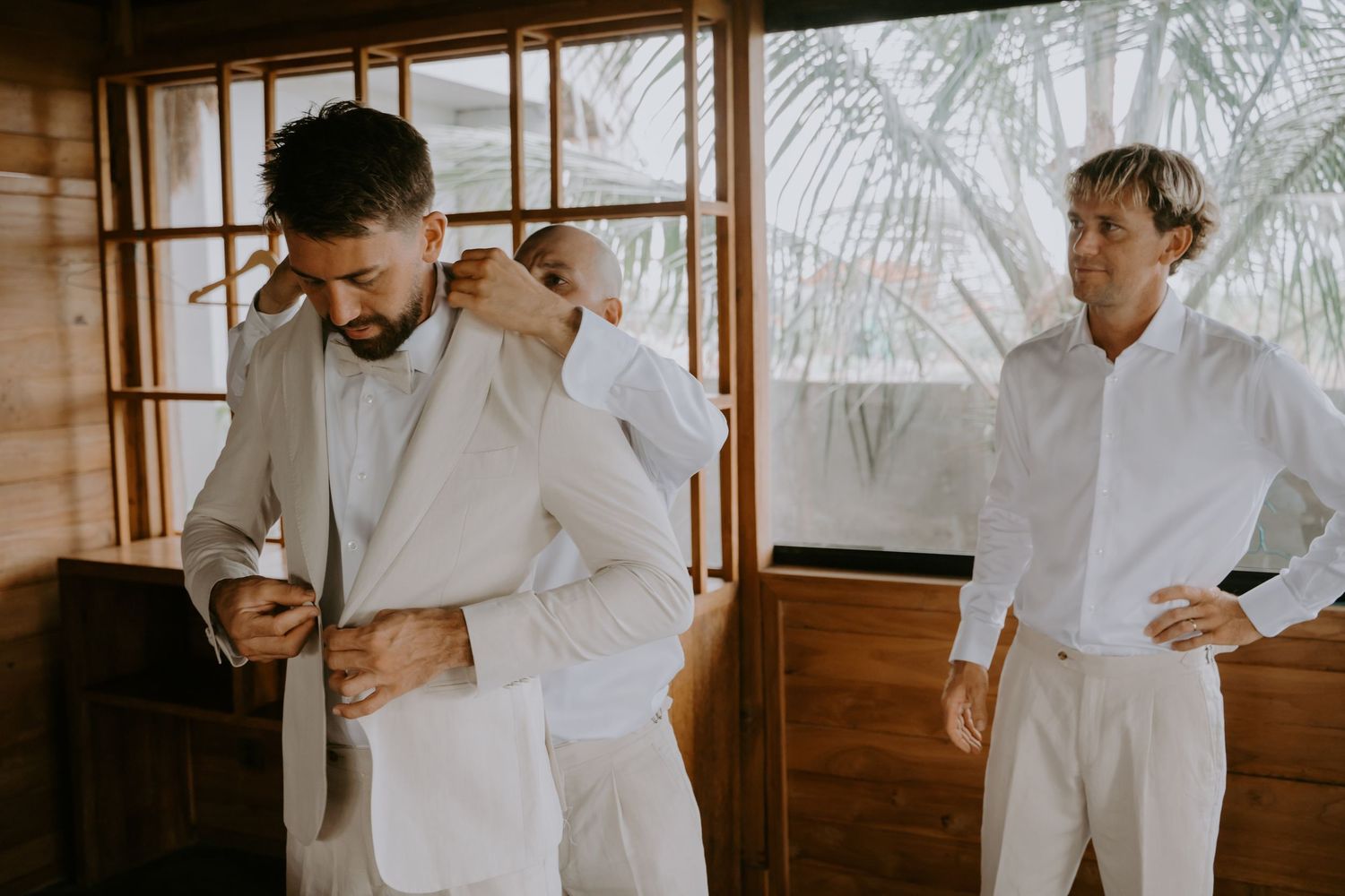 Groom putting on light gray suit jacket assisted by groomsman while another man watches nearby.