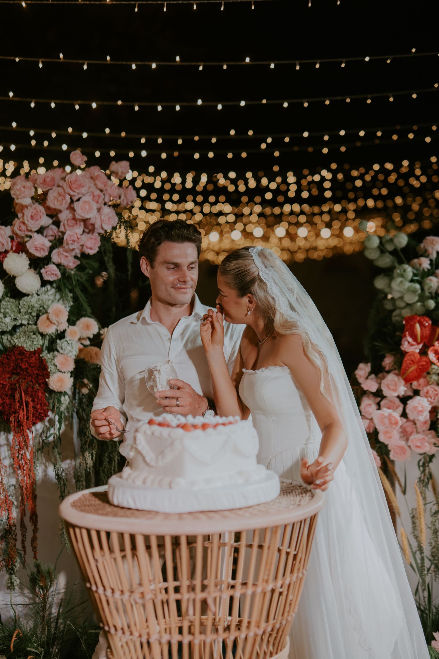 Bride and groom share a joyful moment cutting their strawberry-topped wedding cake under warm string lights.