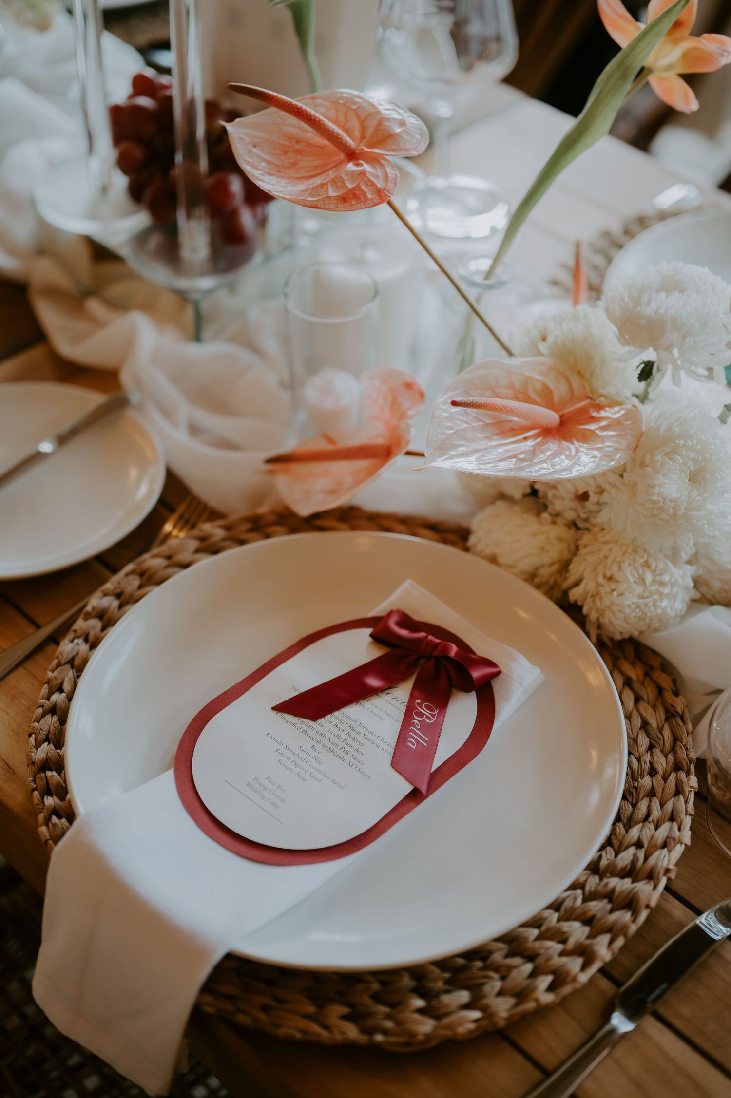 Elegant wedding place setting with woven charger, white plate, burgundy menu card, and tropical floral centerpiece.