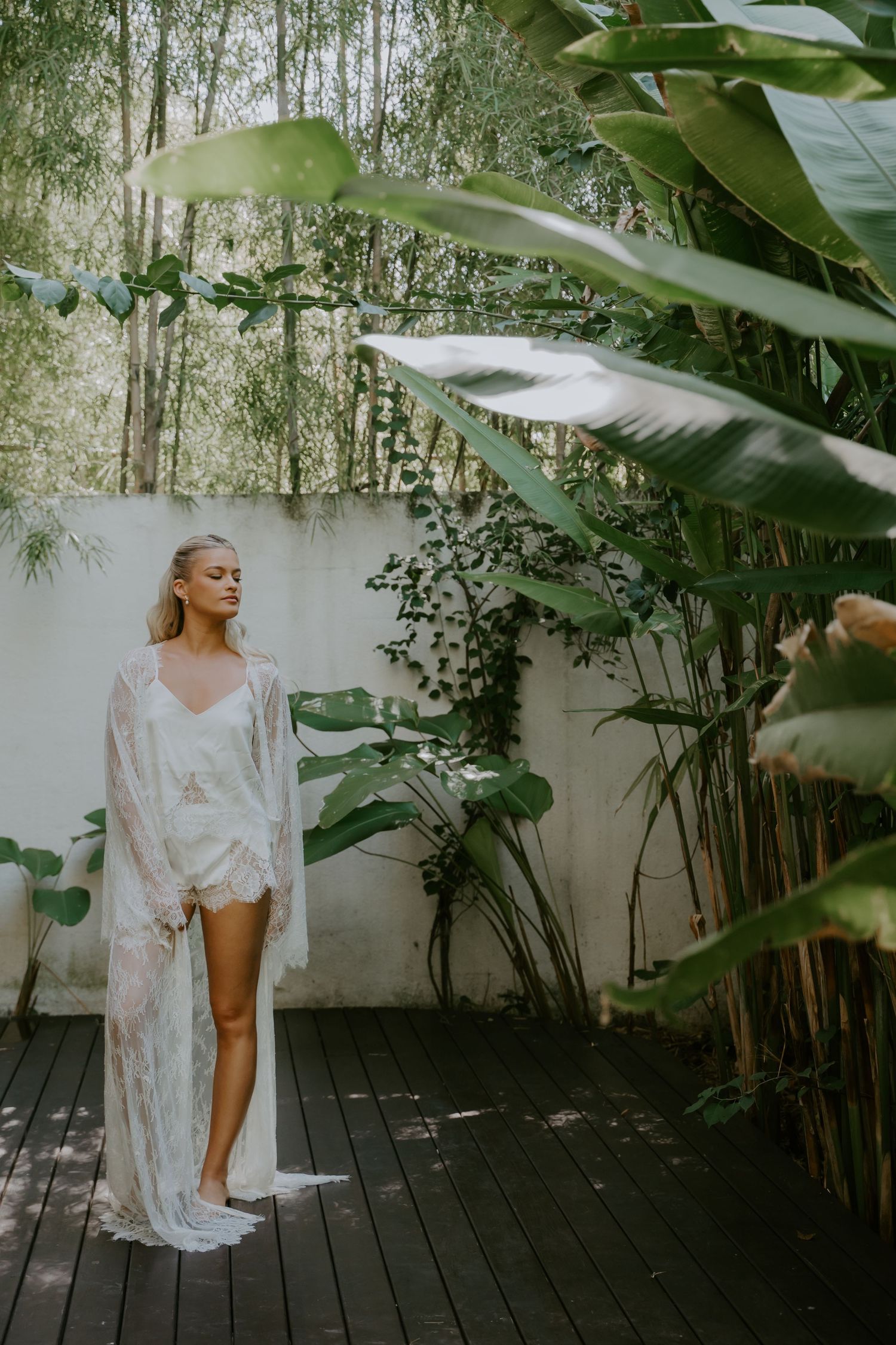Bride in white lace robe standing barefoot on wooden deck surrounded by tropical plants and white wall.