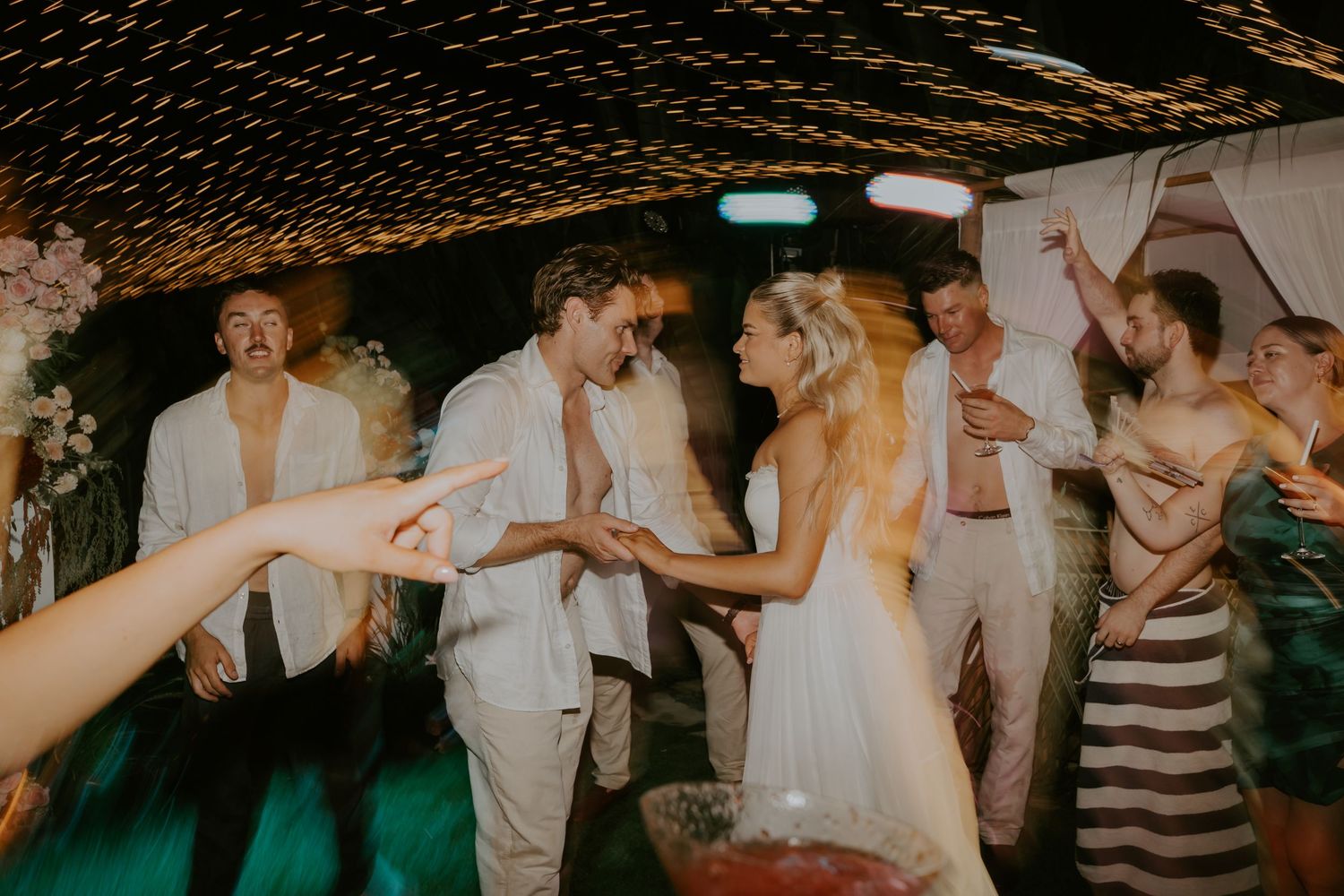 Bride and groom laugh together on outdoor dance floor surrounded by celebrating guests at night reception.