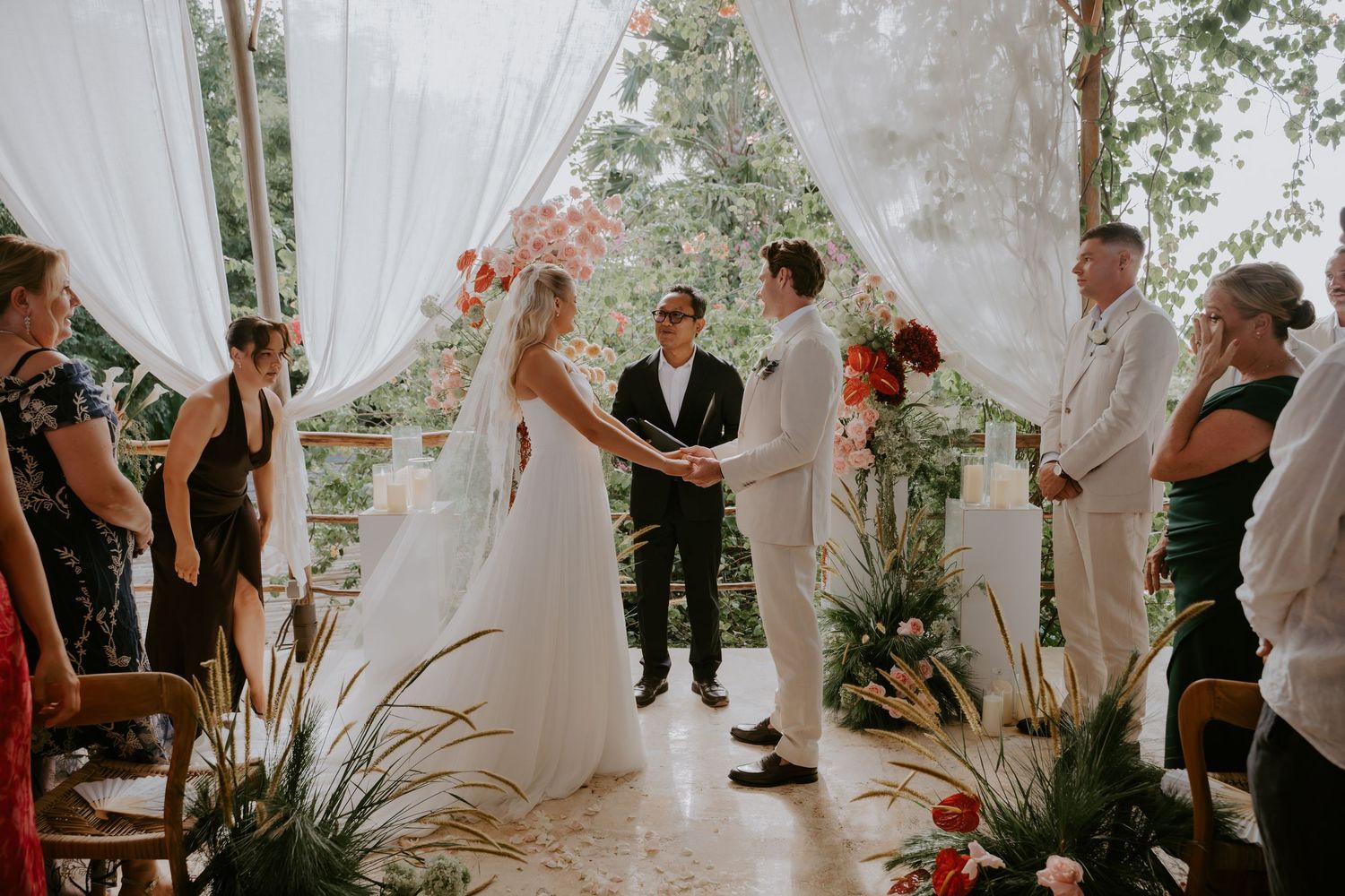 Bride and groom exchange vows at a tropical outdoor ceremony beneath white draped fabric altar with guests.