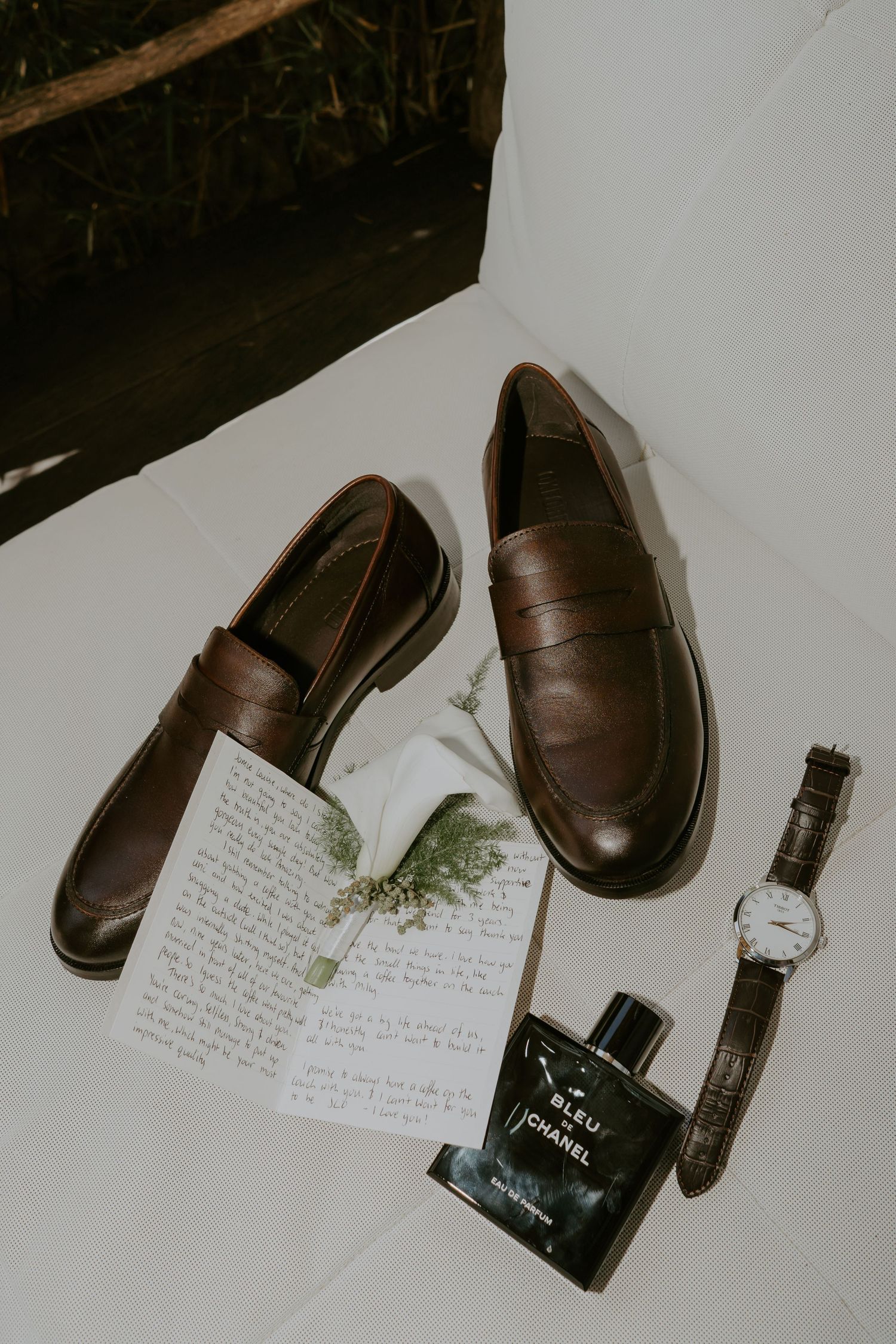 Brown leather penny loafers, Bleu de Chanel cologne, a handwritten letter, watch, and floral sprig arranged as groom wedding details.