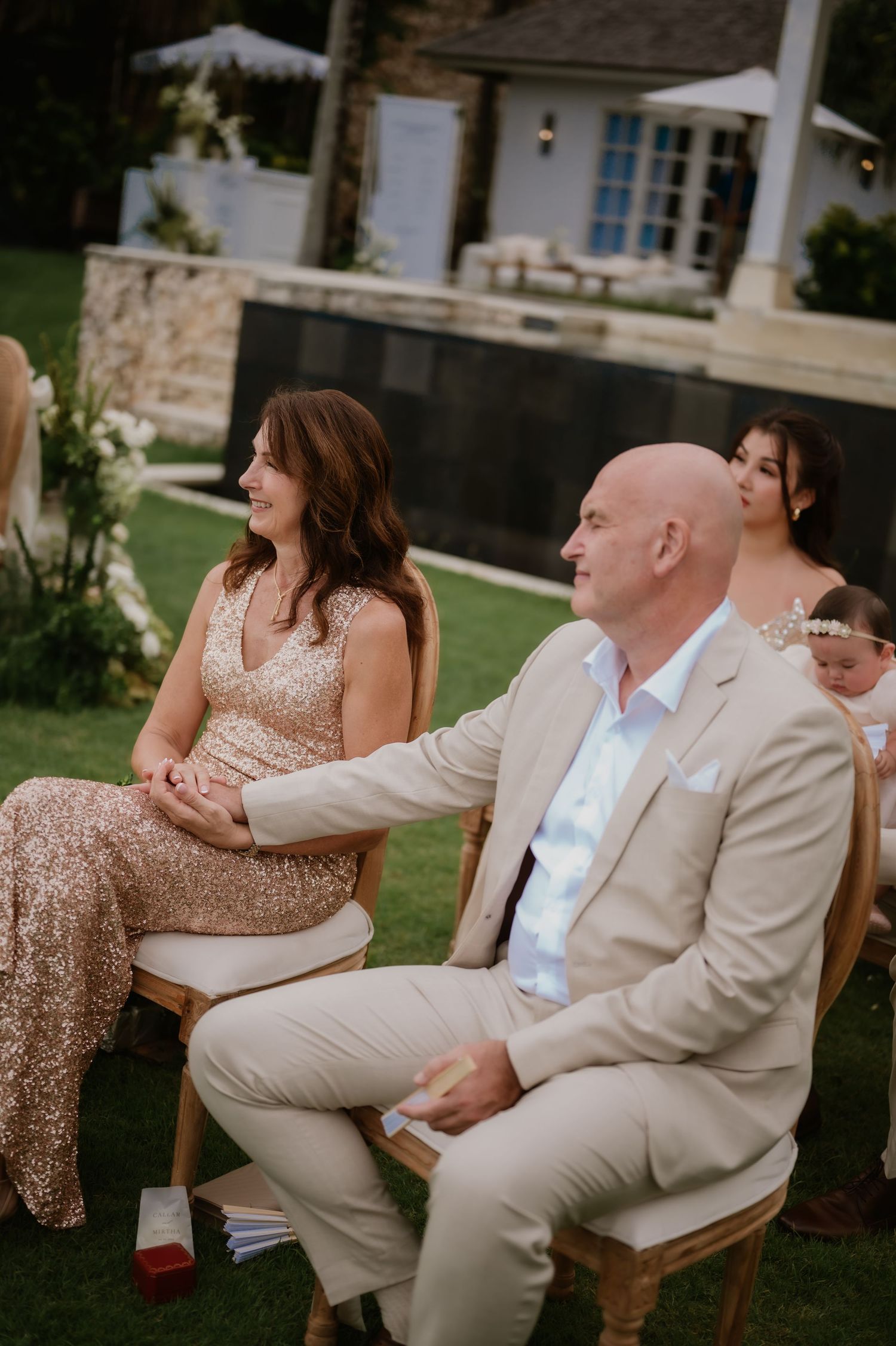 Guests seated outdoors at an elegant wedding ceremony, man in beige suit holds woman's hand in sequin dress.