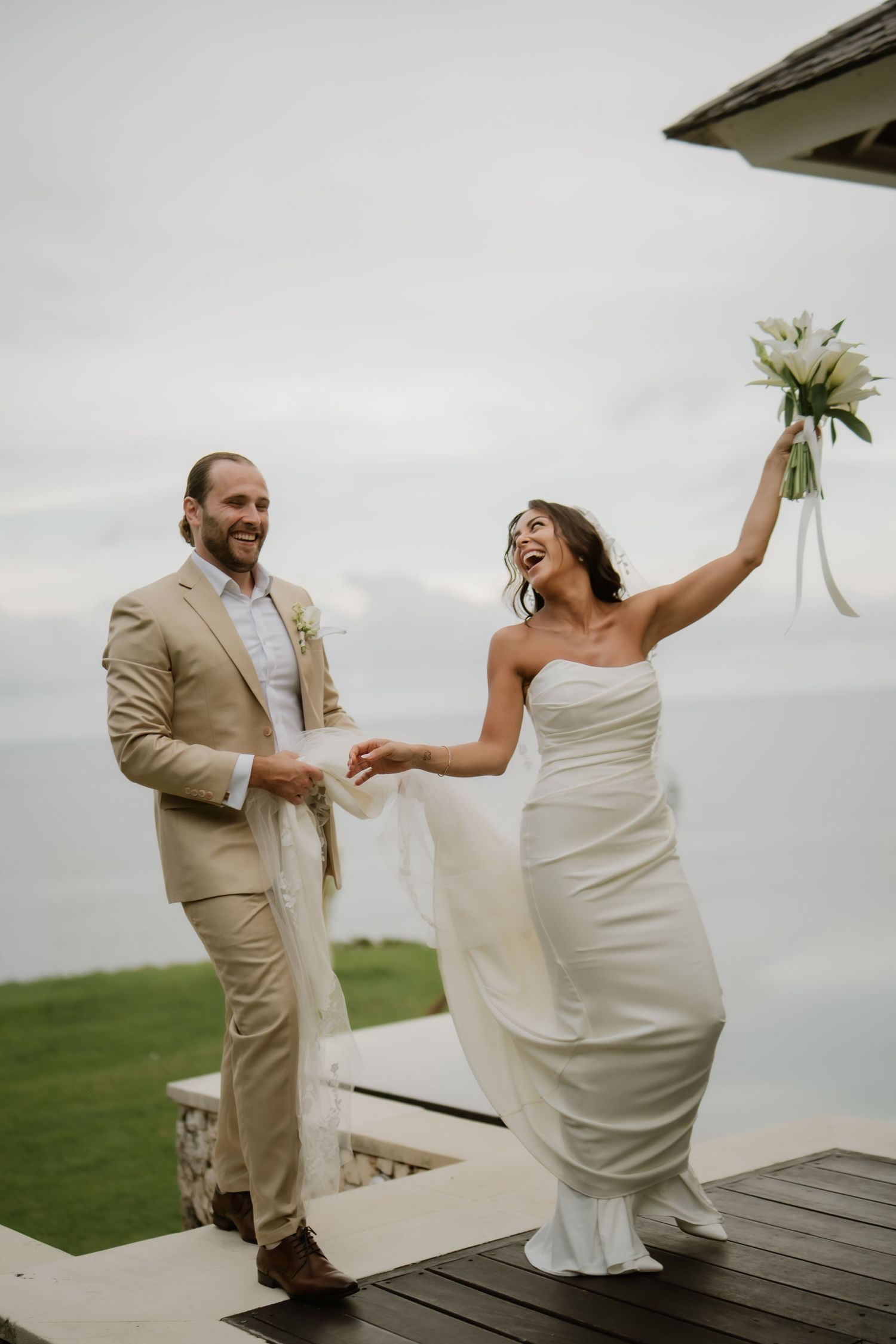 Joyful bride raises bouquet while groom holds her hand at outdoor seaside wedding venue.