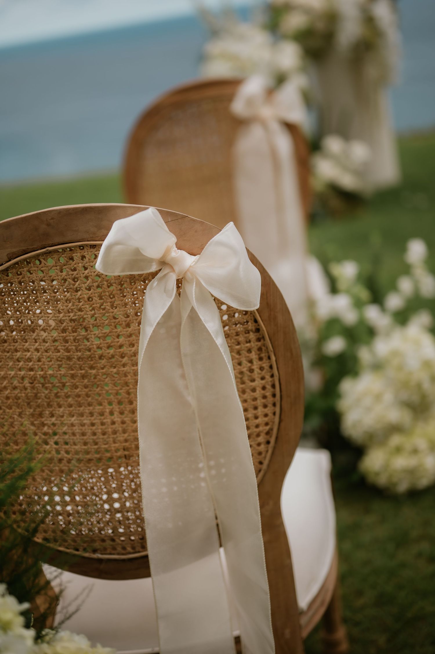 Close-up of a rattan wedding chair with an ivory silk bow tied to the back overlooking the ocean.