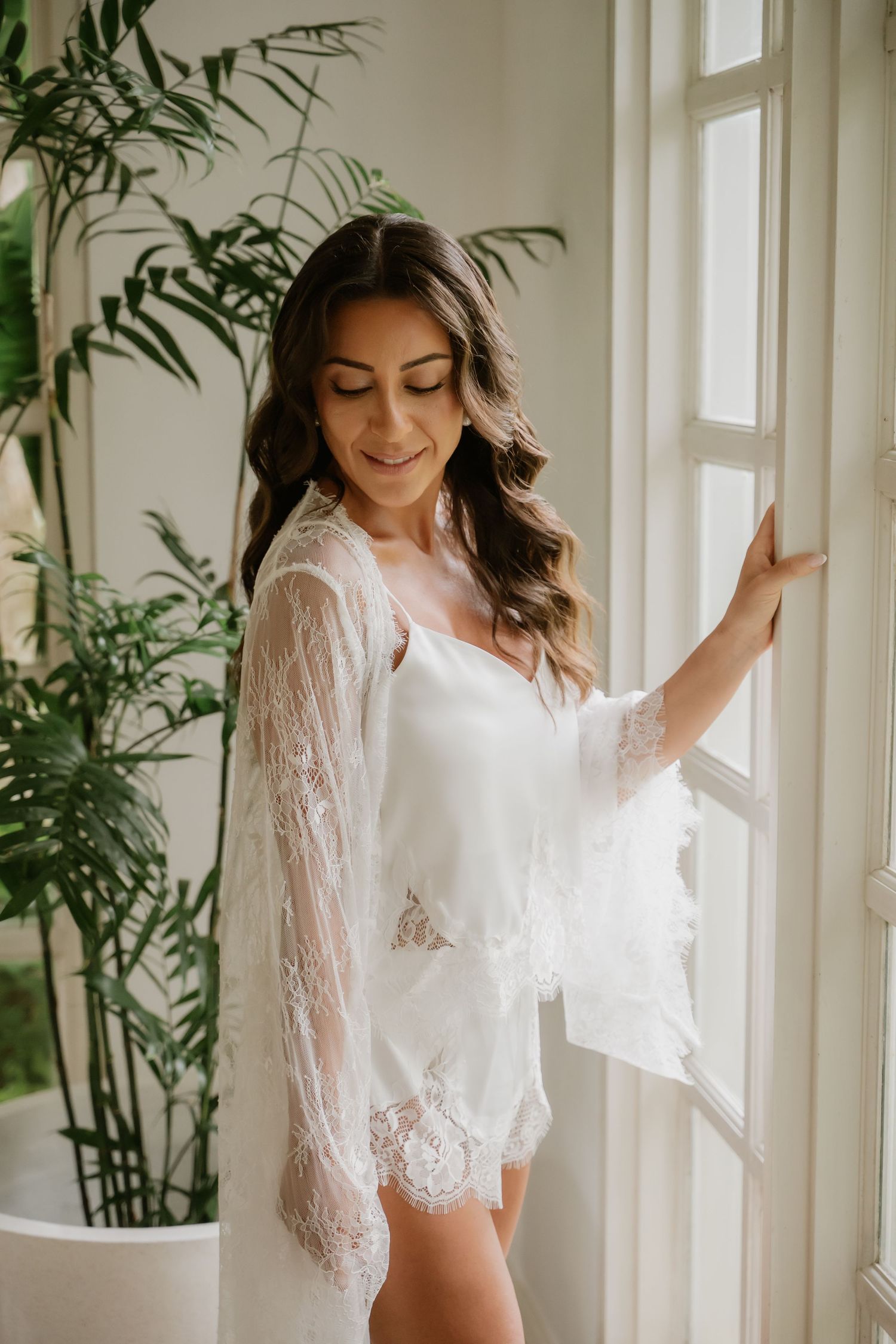 Beautiful bride in white lace robe smiling softly while standing by a sunlit window with tropical plants.