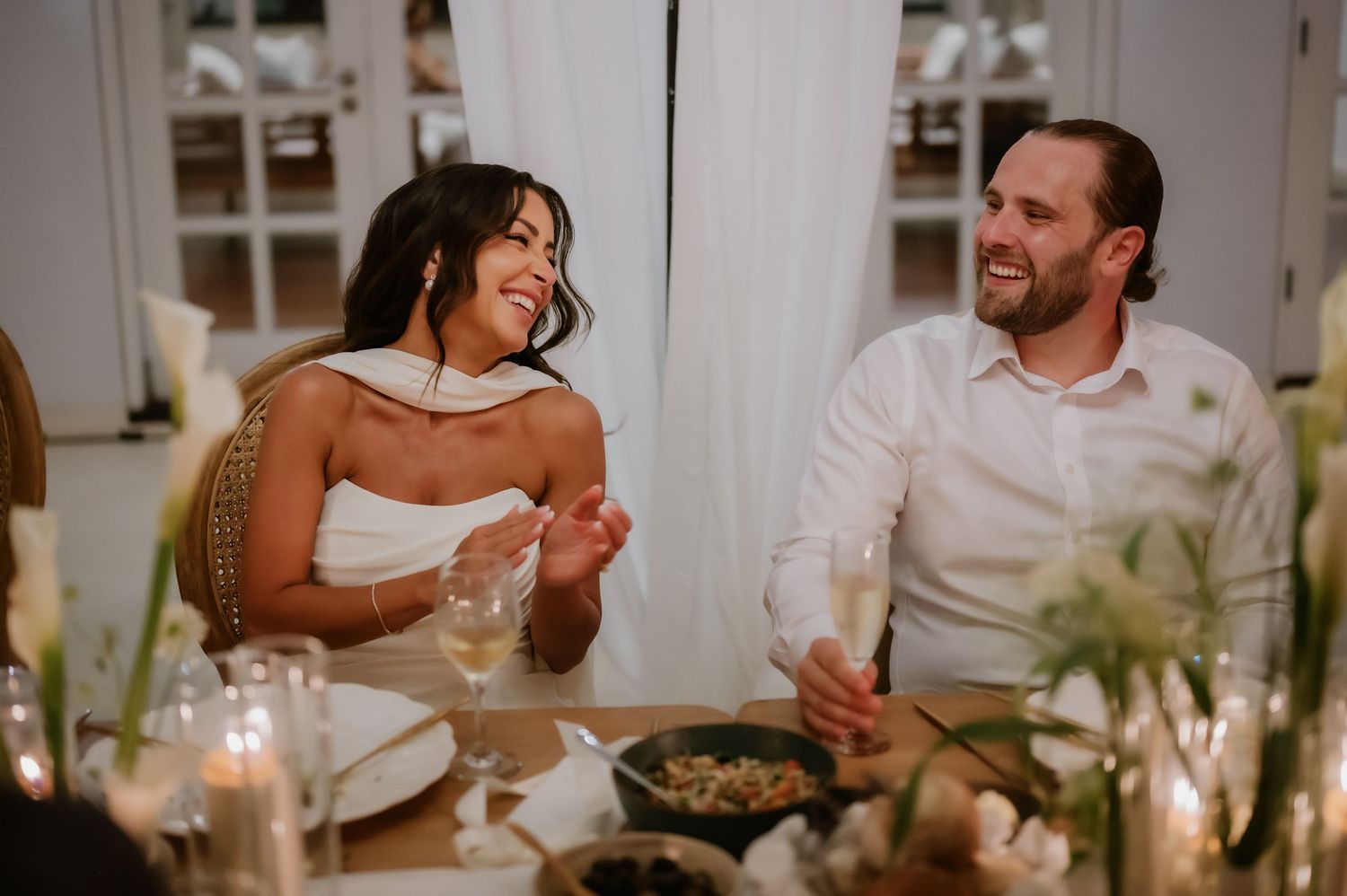 Bride and groom laugh joyfully together at candlelit wedding reception dinner table.