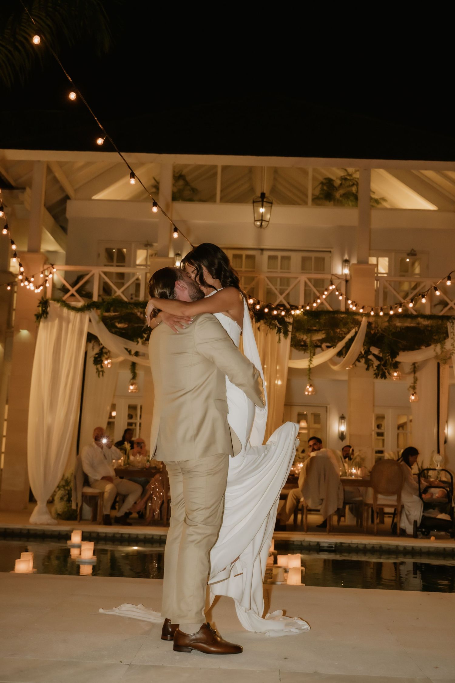 Groom lifting bride in white wedding gown for romantic kiss beside glowing pool under string lights.