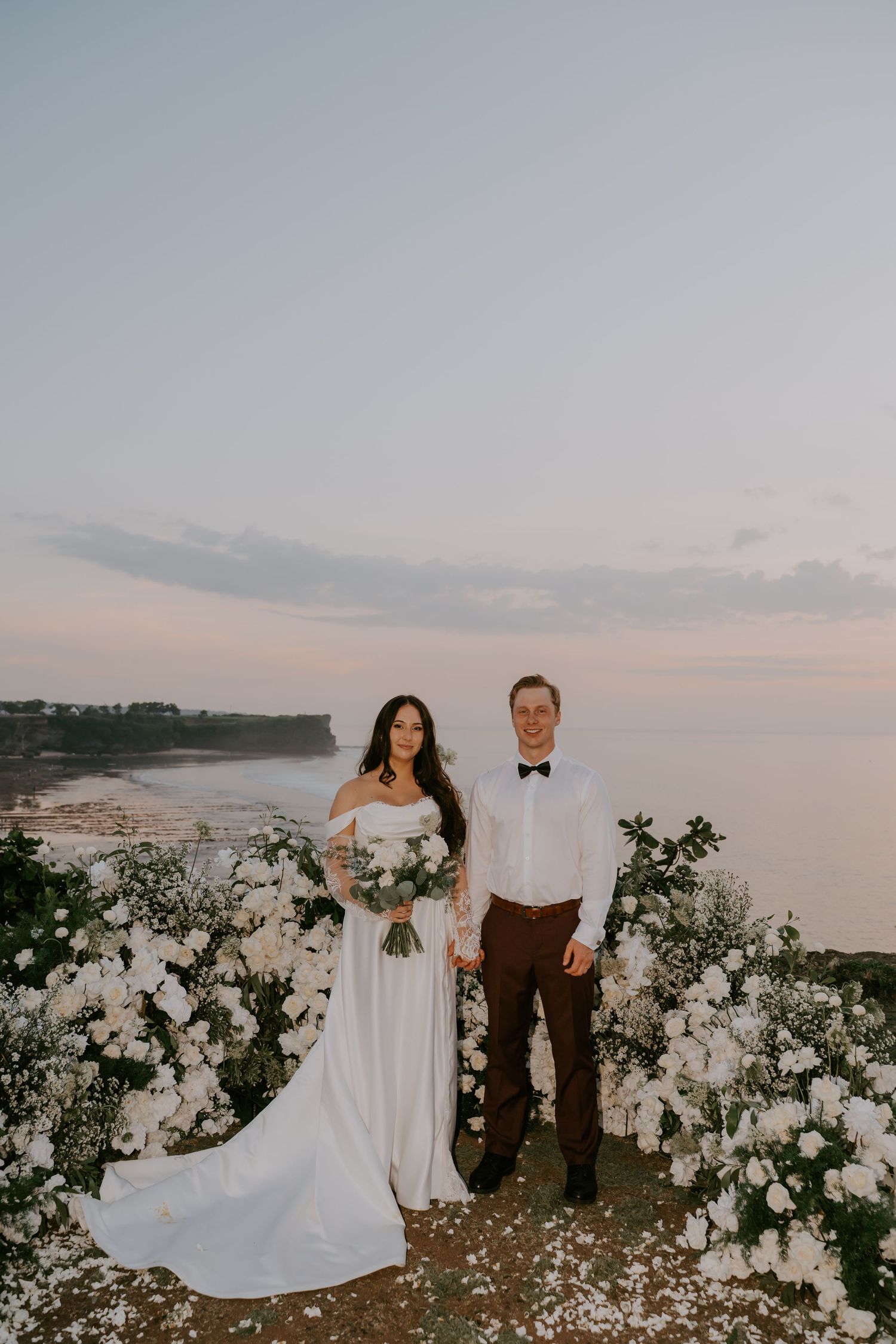 Bride and groom pose together at sunset with white floral arch and ocean view behind them.