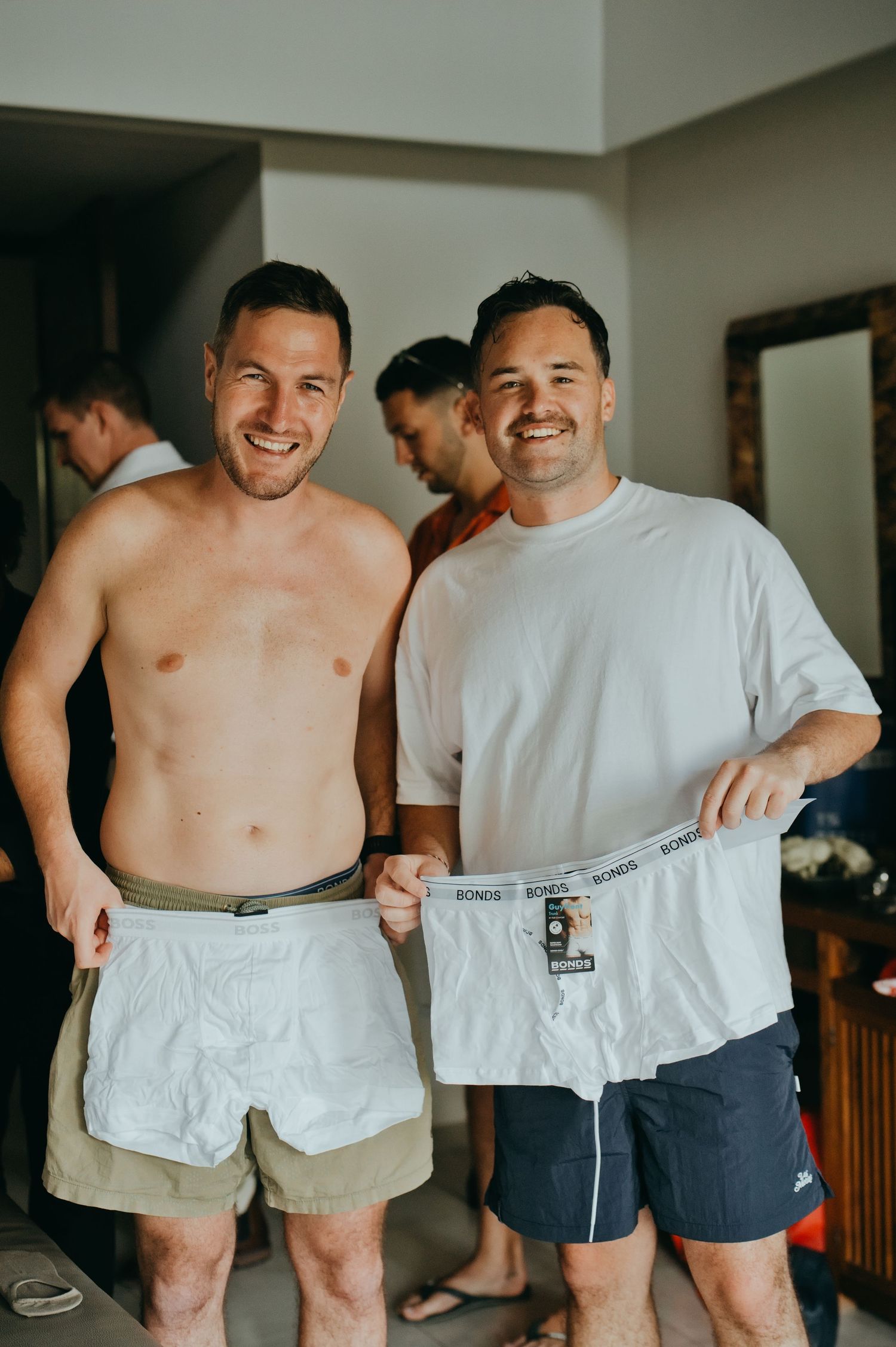 Two smiling groomsmen holding Bonds underwear during wedding preparations indoors.