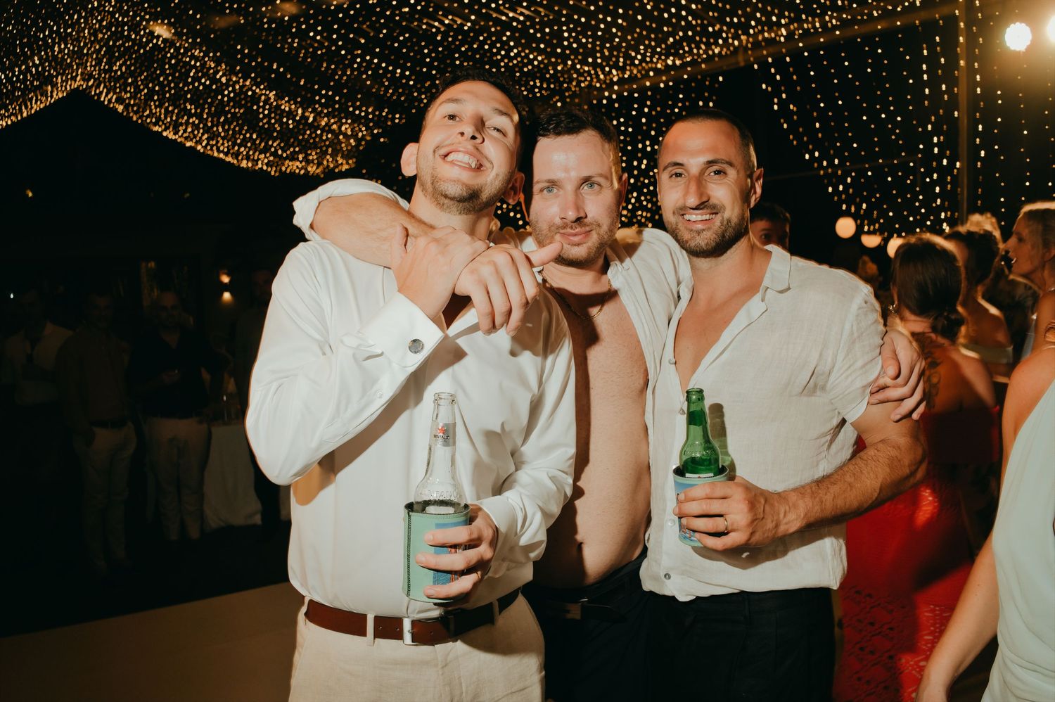 Three groosmen celebrating at nighttime wedding reception under string lights while holding beer bottles and smiling.