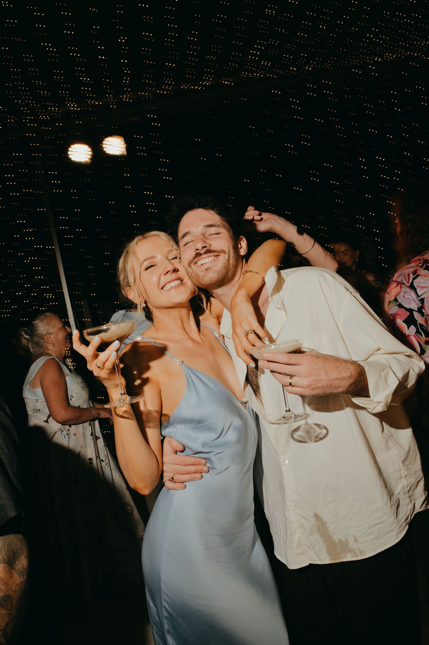 Bridesmaid in a blue satin dress and male guest smile and hold cocktail glasses at a wedding reception.