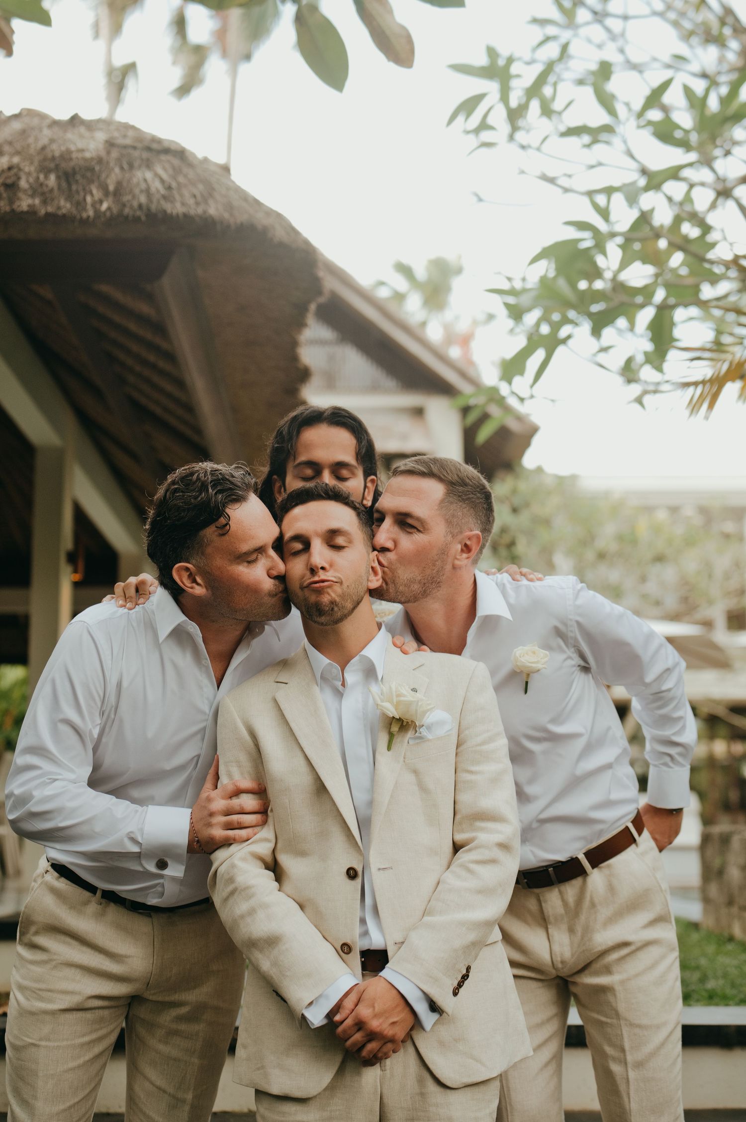 Two groomsmen kissing the groom on the cheeks during a fun wedding day photo at a tropical venue.