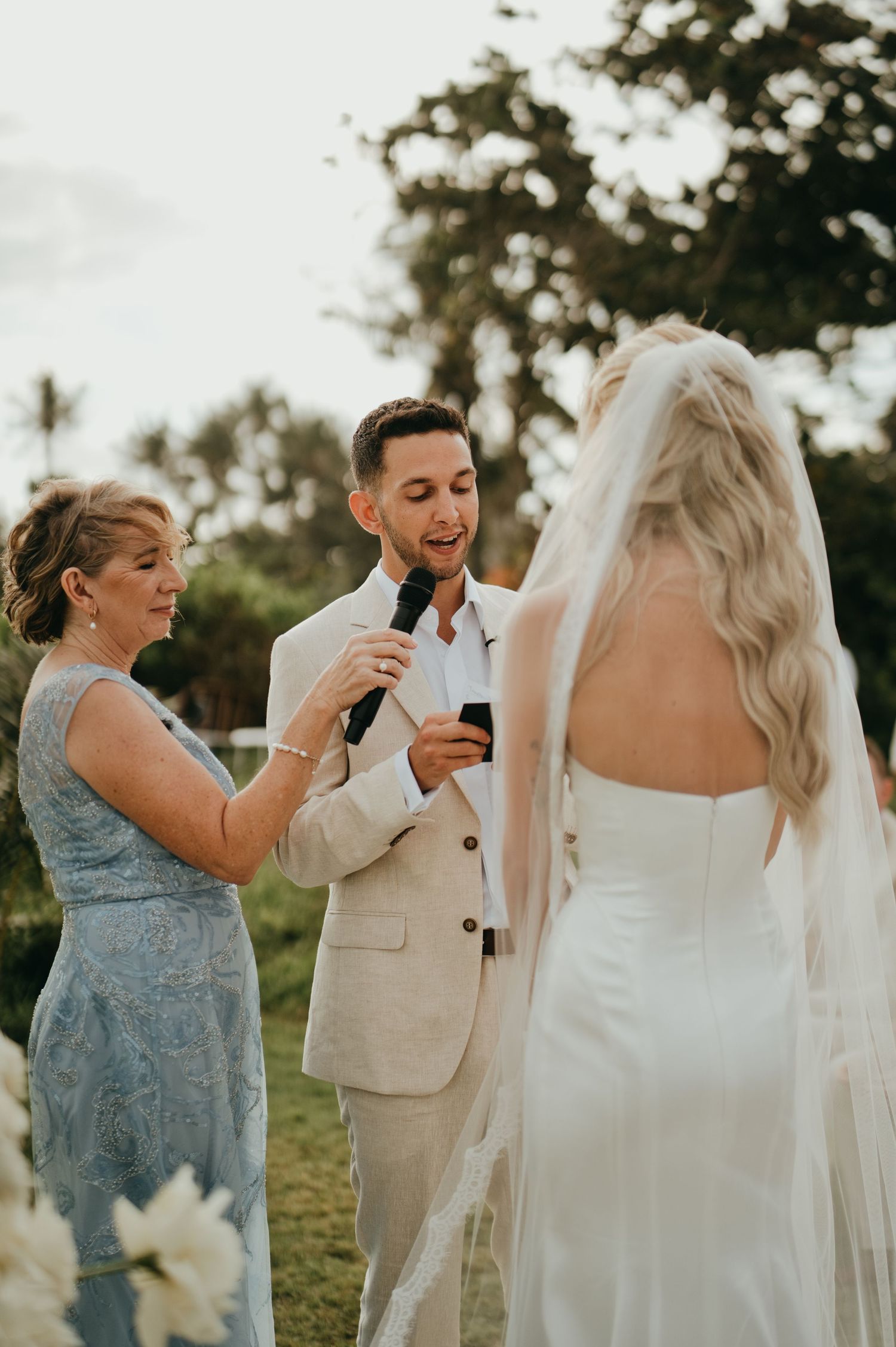 Groom in beige suit reads vows to bride in white backless gown at outdoor ceremony.