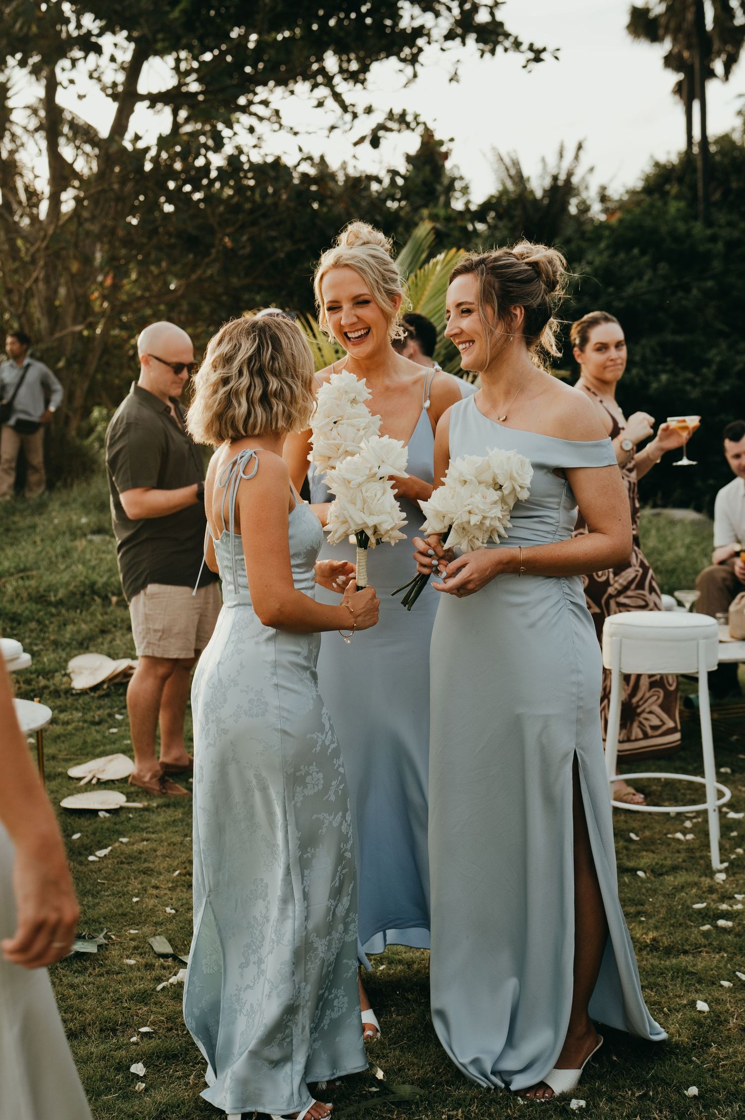 Three bridesmaids in light blue gowns laugh together holding white floral bouquets at outdoor wedding reception.