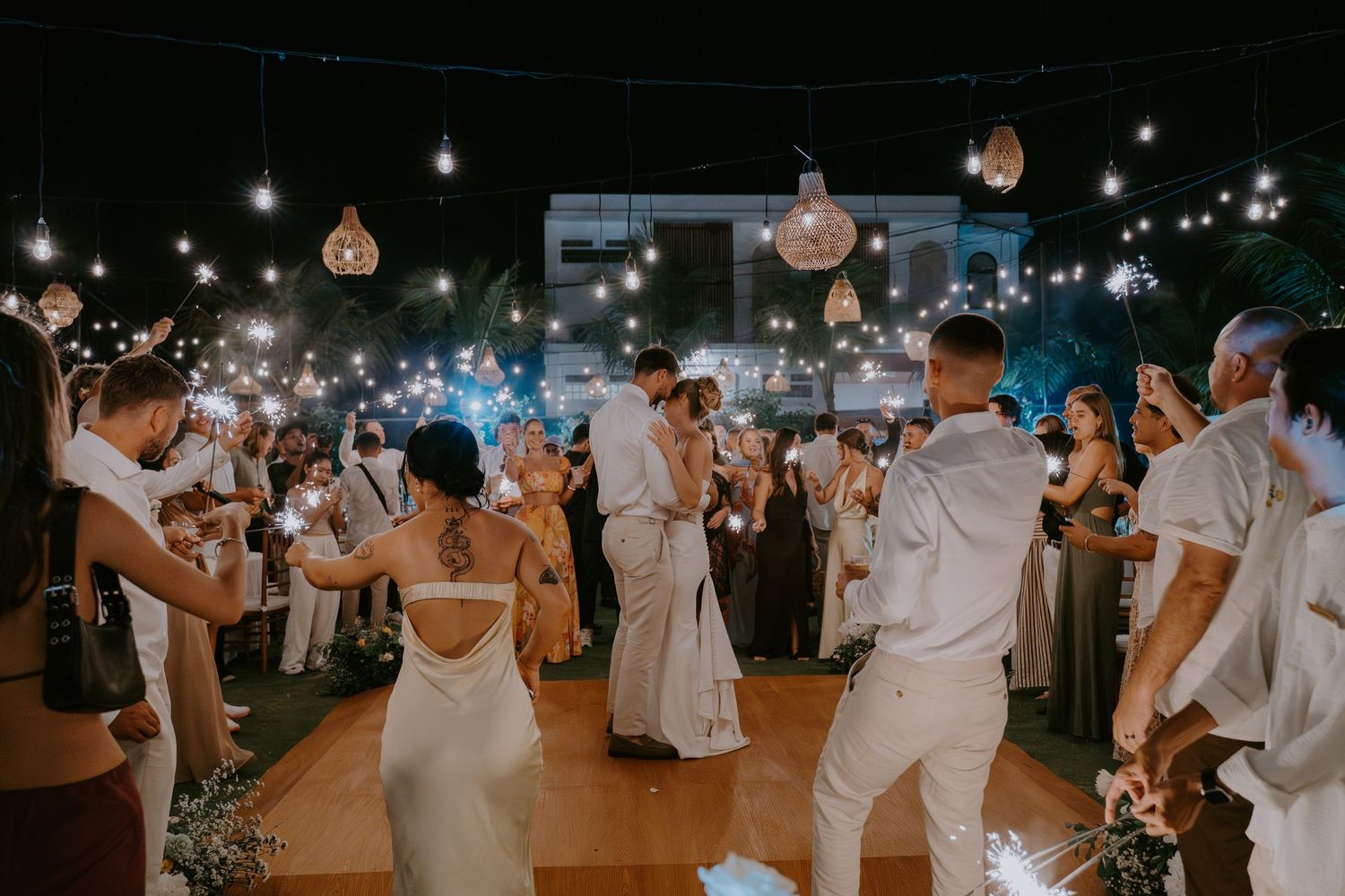 Bride and groom share first dance surrounded by guests holding sparklers at an elegant outdoor wedding reception at night.
