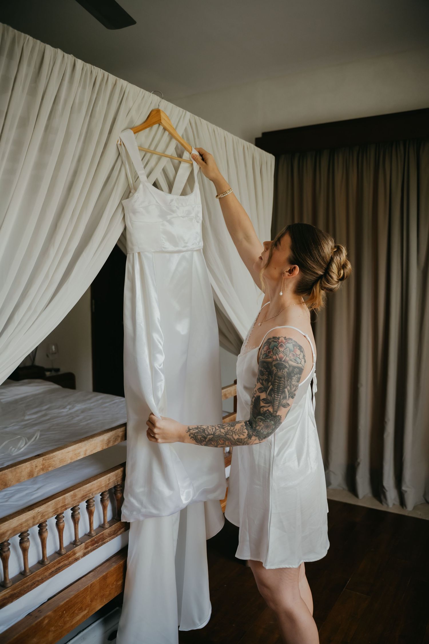 Tattooed bride in white robe admiring her wedding dress hanging on a canopy bed in a hotel room.