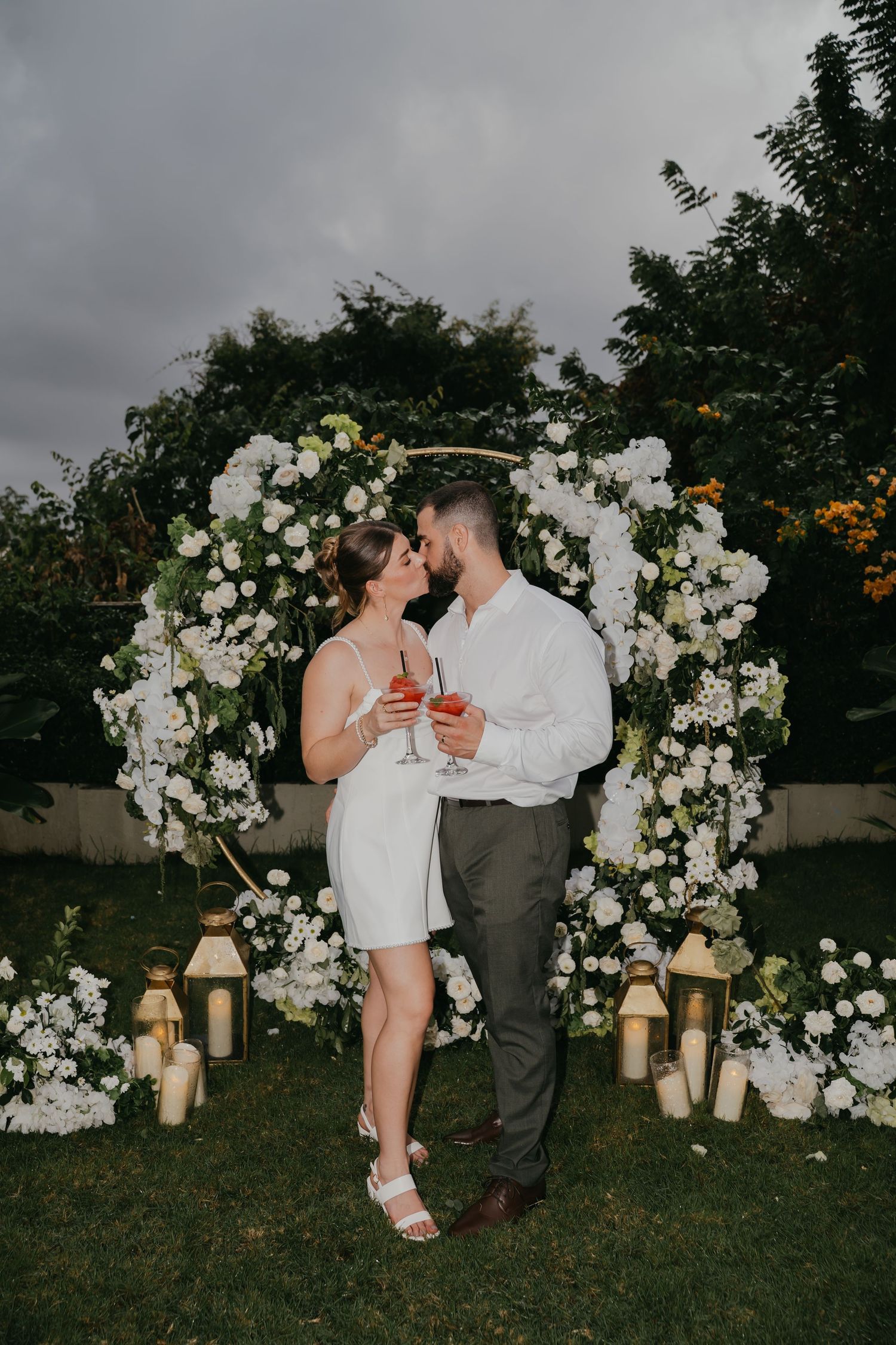 Newlyweds kiss holding red drinks before white floral arch with lanterns at dusk.