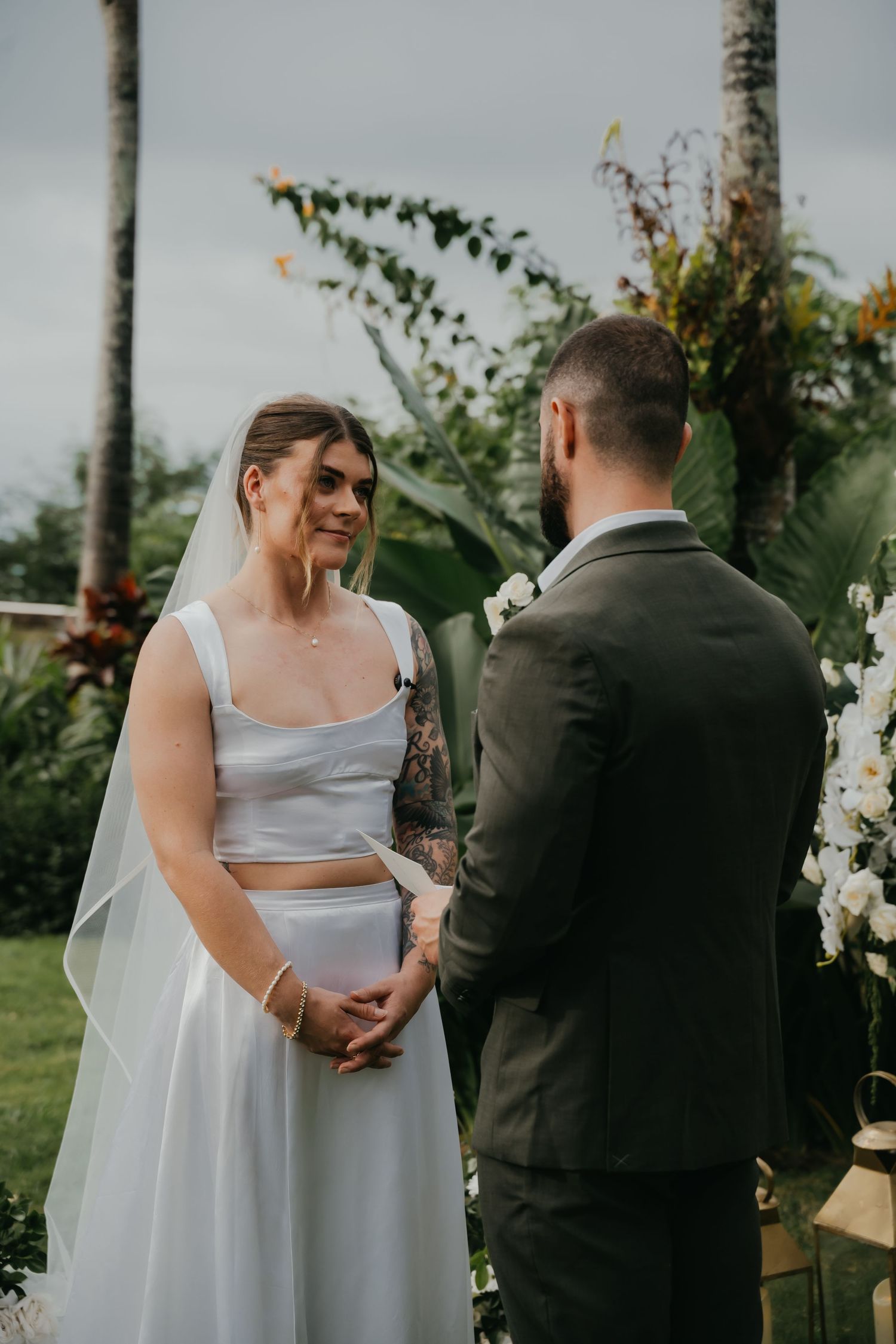 A smiling bride in a two-piece white gown exchanges vows with a groom at an outdoor ceremony.