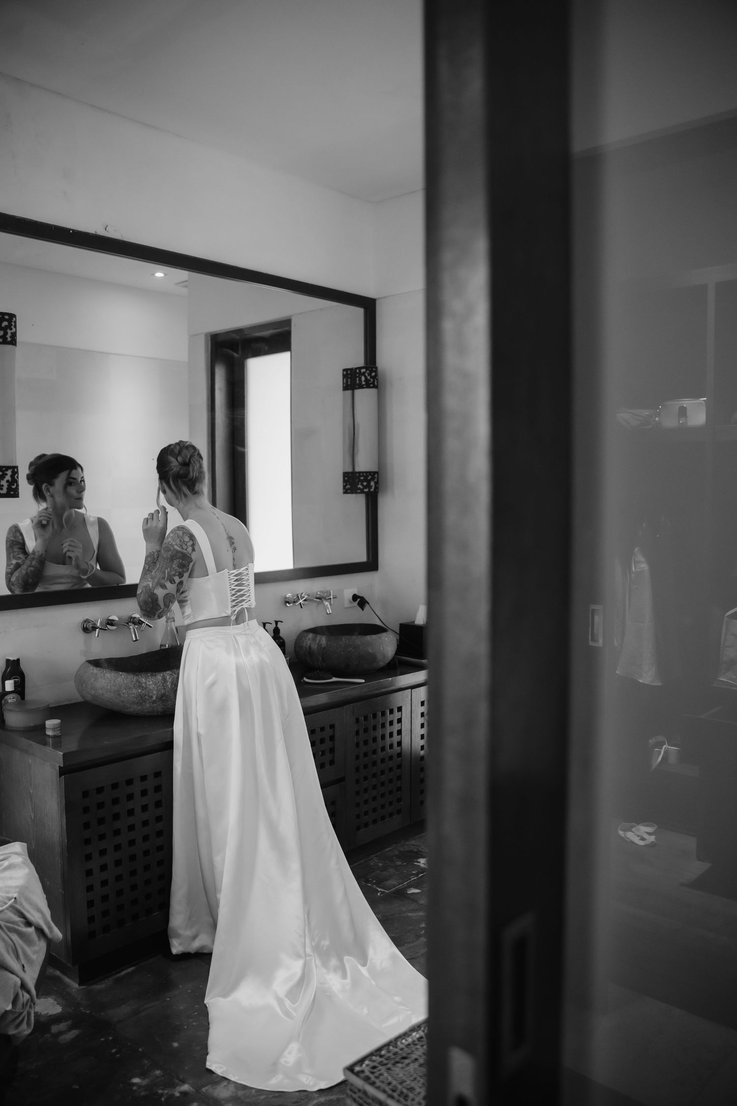 Bride in a white gown with lace back stands before bathroom mirror in black and white wedding photo.