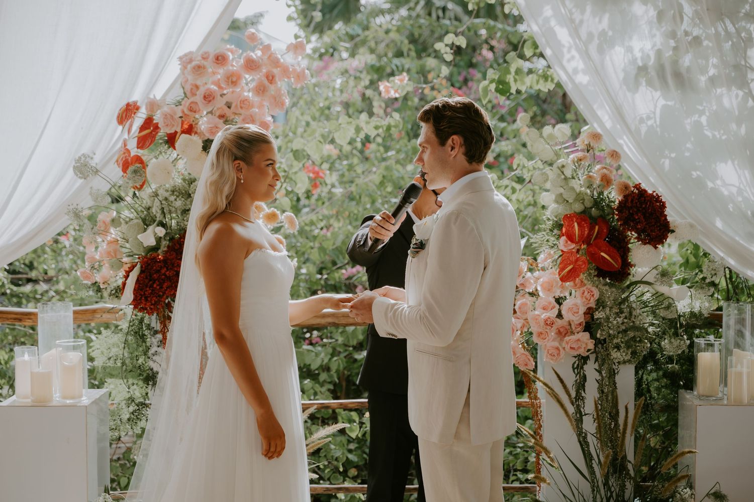 Groom reads wedding vows to bride beneath a white draped arch with pink floral decor.