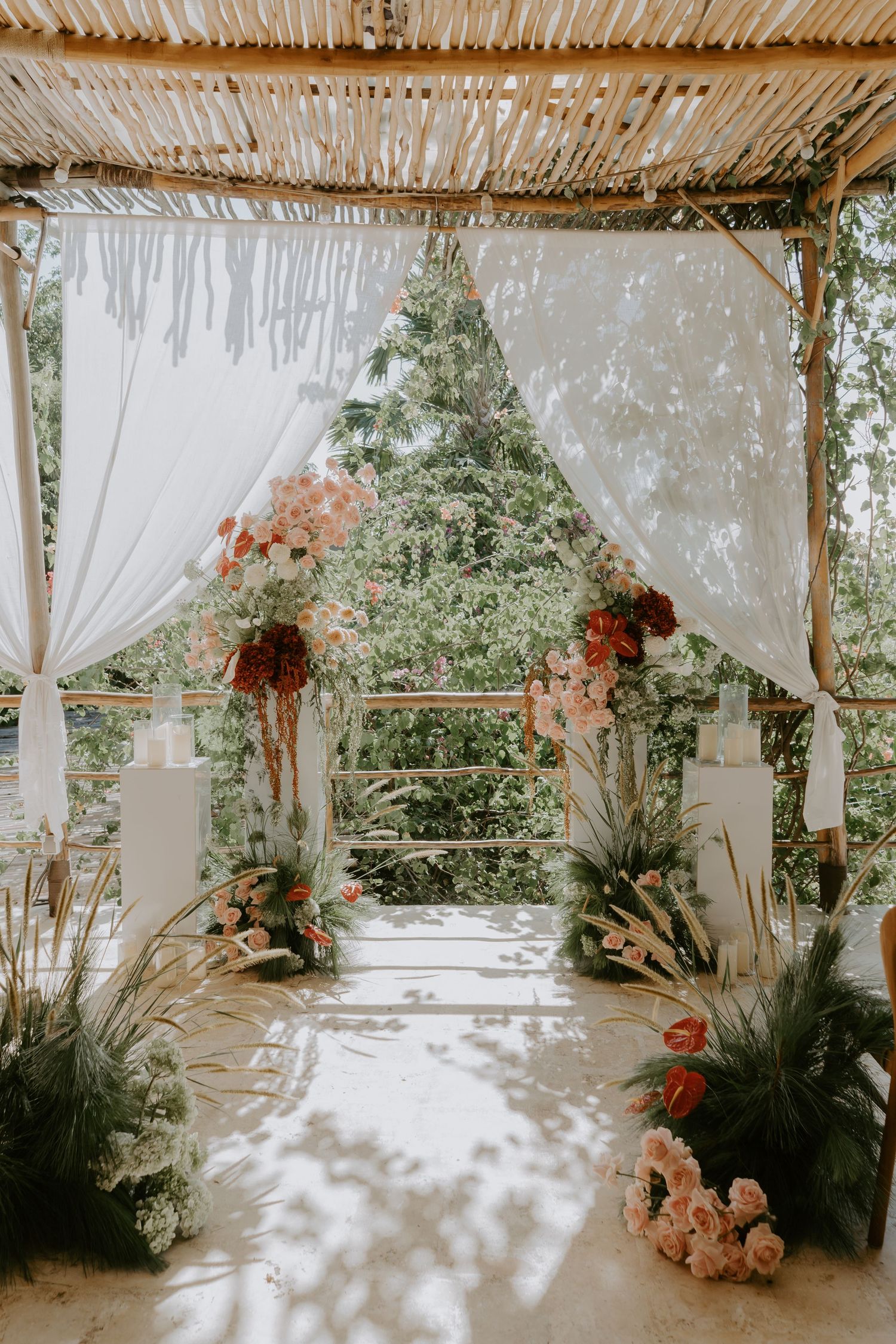 Elegant outdoor wedding arch with white drapes, pink and red floral arrangements under bamboo roof.