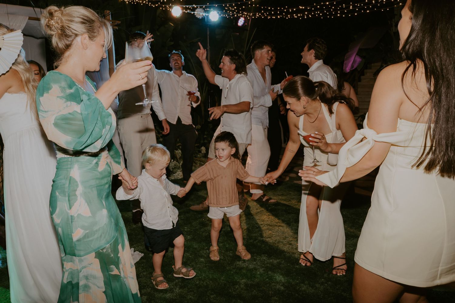 Children and wedding guests dance together outdoors under string lights during a festive evening wedding reception.
