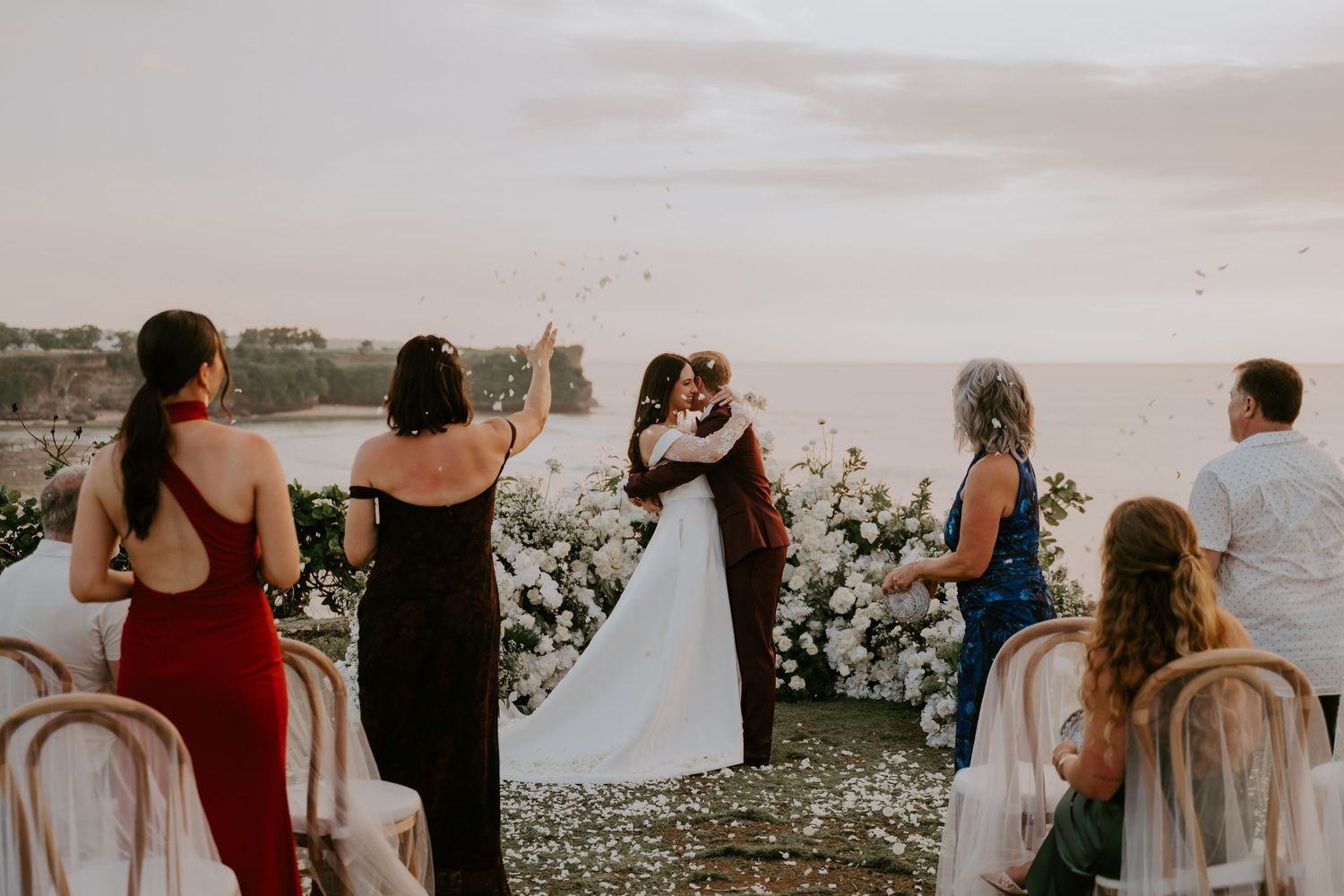 Wedding guests toss petals as couple embraces at cliffside ceremony with ocean views at sunset.