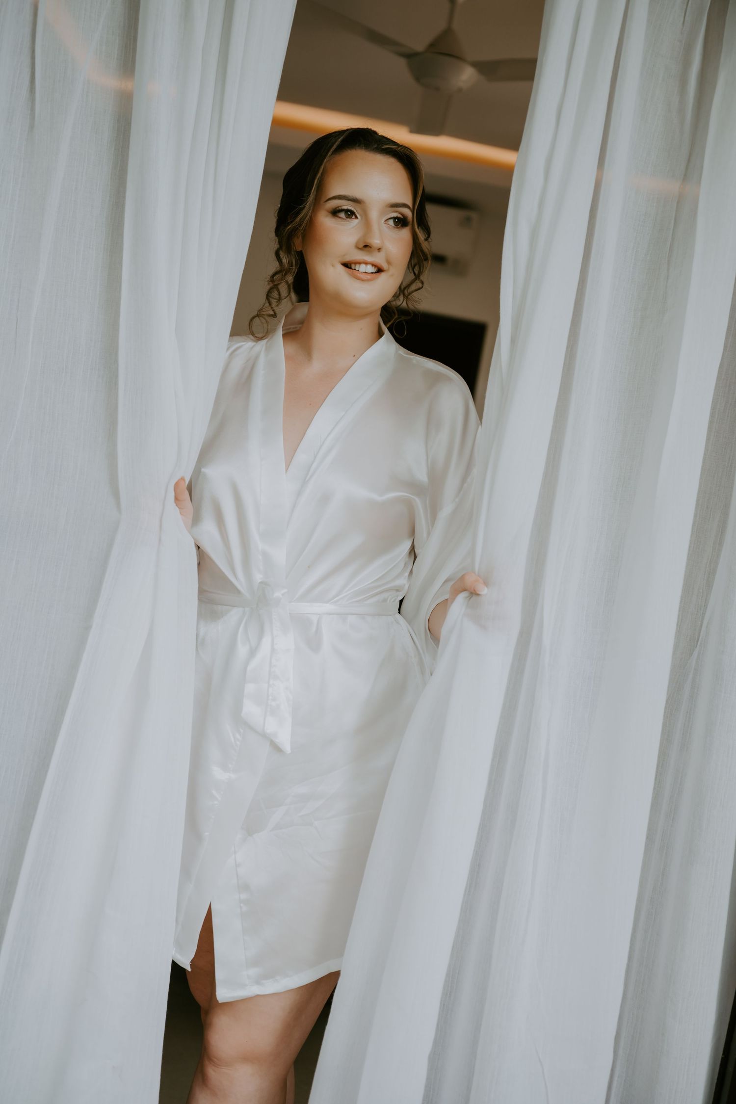 Bride in white satin robe standing between sheer curtains, smiling softly on her wedding morning.