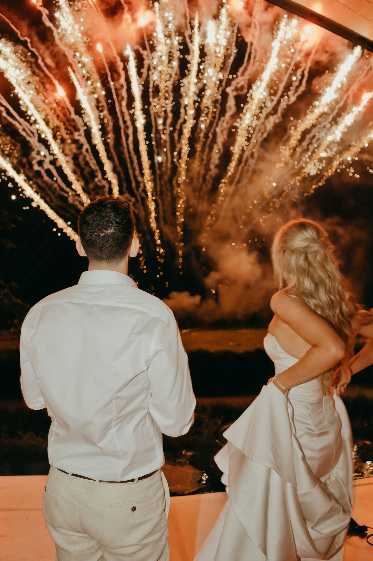 Bride and groom watch stunning fireworks display together at romantic outdoor wedding reception at night.