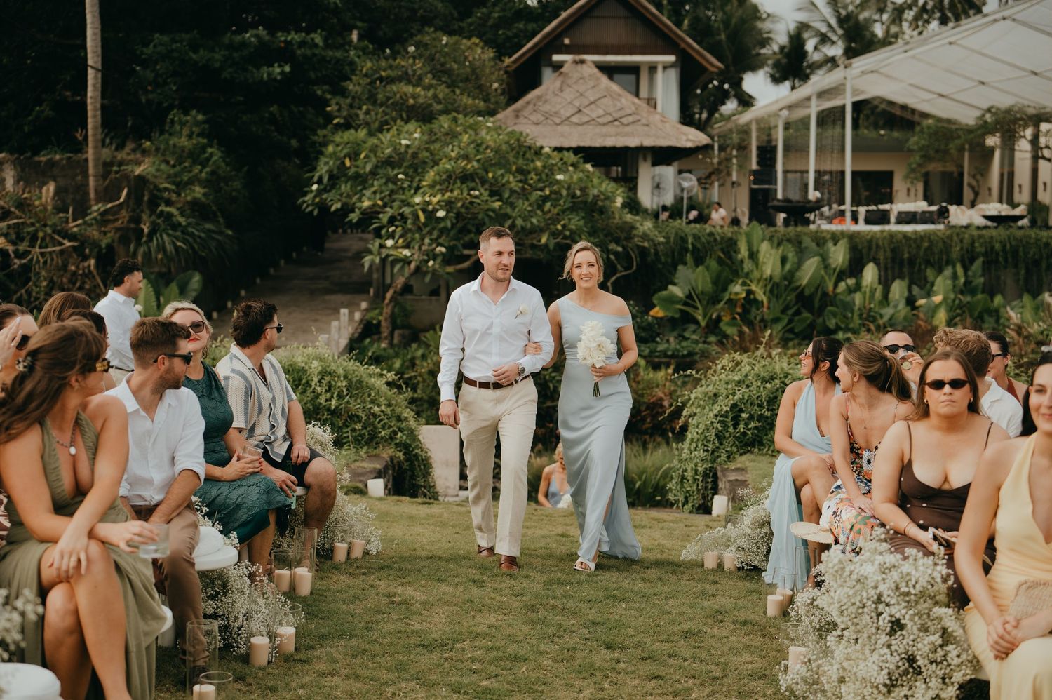 Wedding guests walking down grass aisle at outdoor ceremony with ocean backdrop.