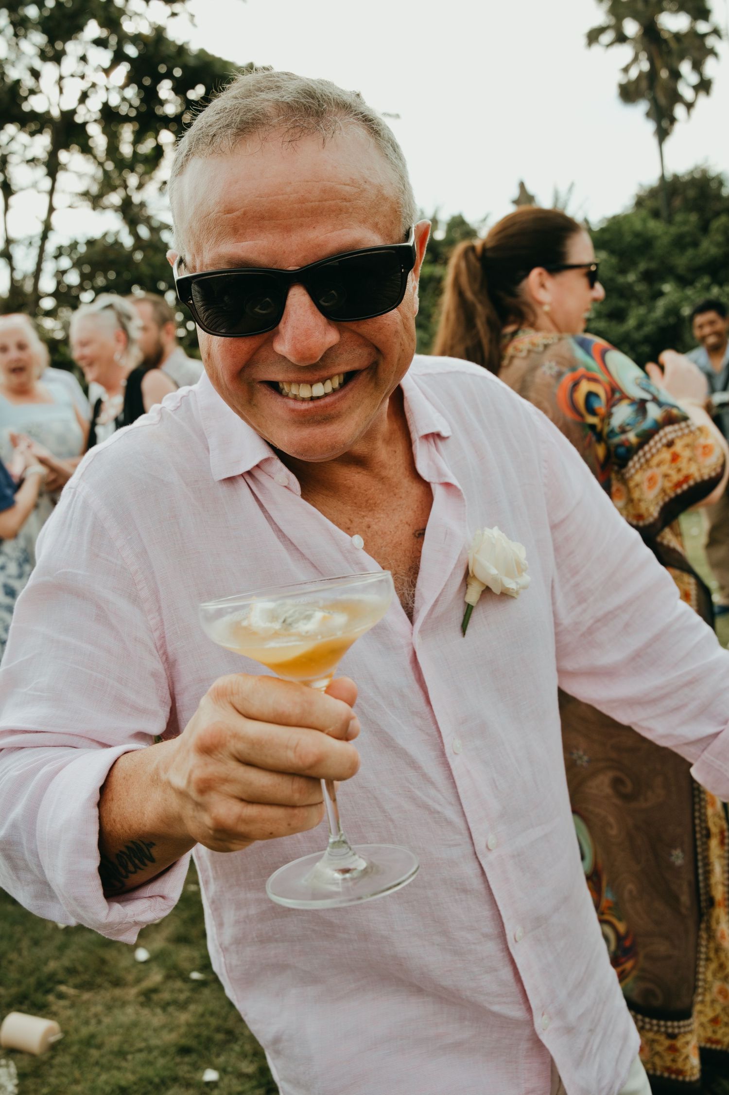 Smiling older man in pink shirt raises a cocktail glass at an outdoor wedding reception.
