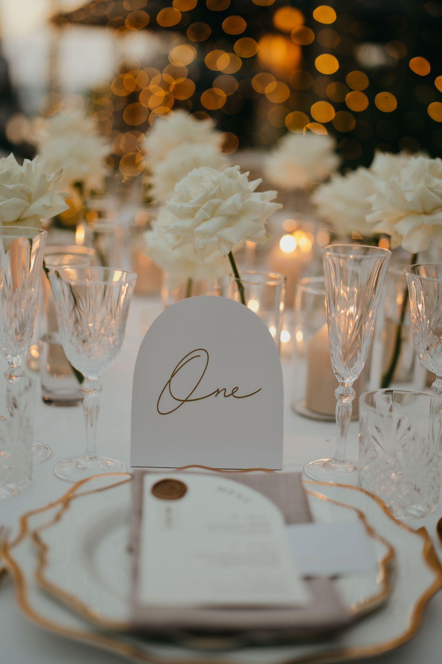 Elegant wedding table setting with white floral centerpieces, crystal glassware, and gold-trimmed arch table number.
