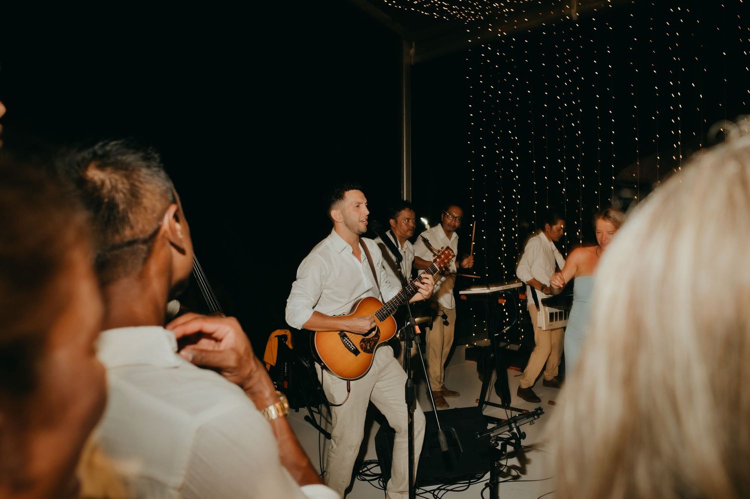Live wedding band guitarist performs energetically for guests at a twinkling fairy-lit outdoor reception.