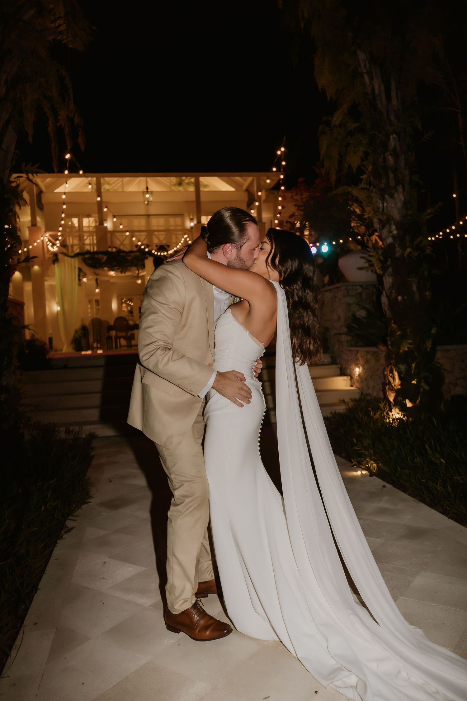 Bride and groom kissing at night with string lights and tropical venue glowing behind them.
