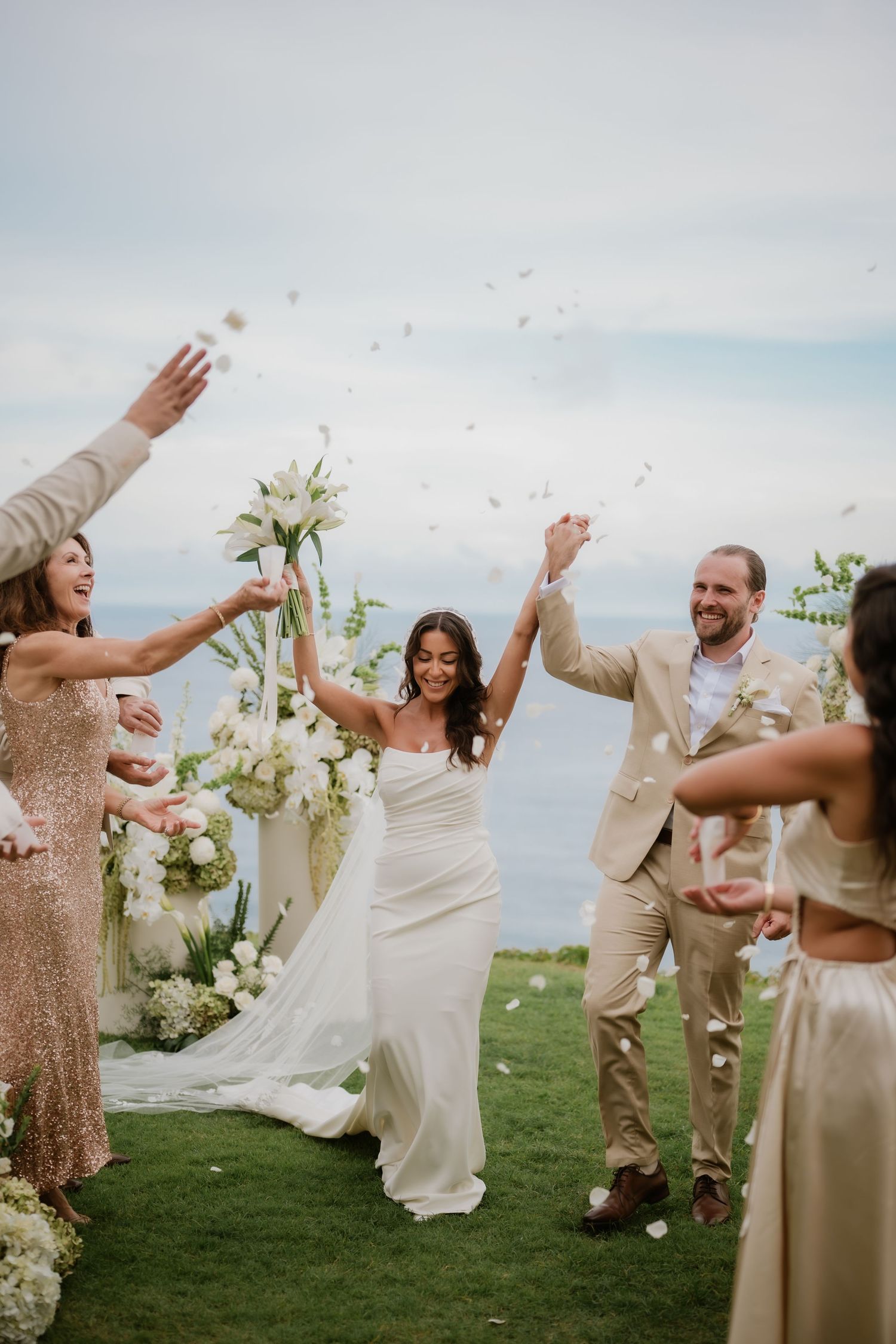 Bride and groom celebrate recessional as guests toss white flower petals outdoors by the sea.