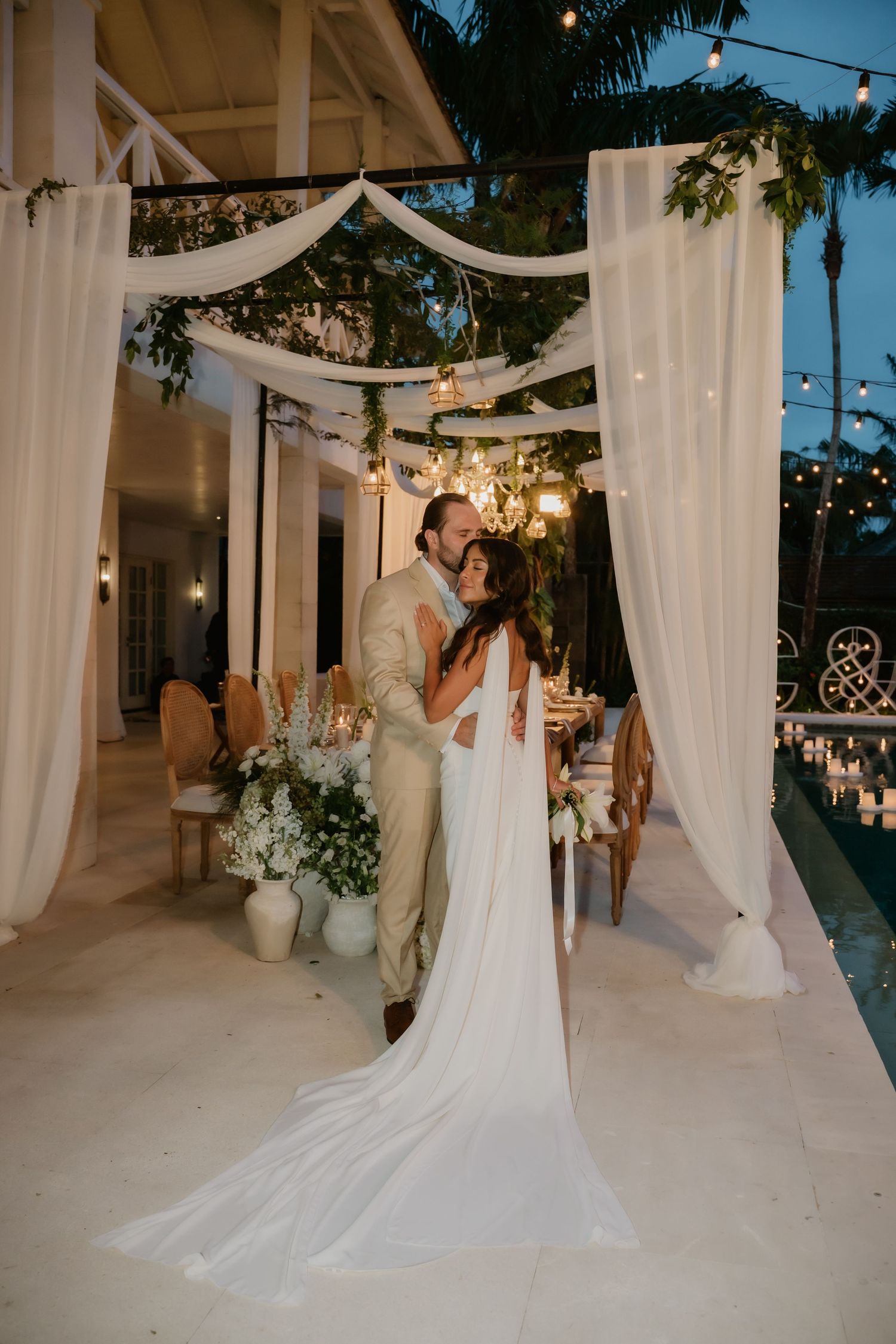 Bride and groom share a romantic kiss under white draped canopy with string lights at elegant outdoor wedding reception.