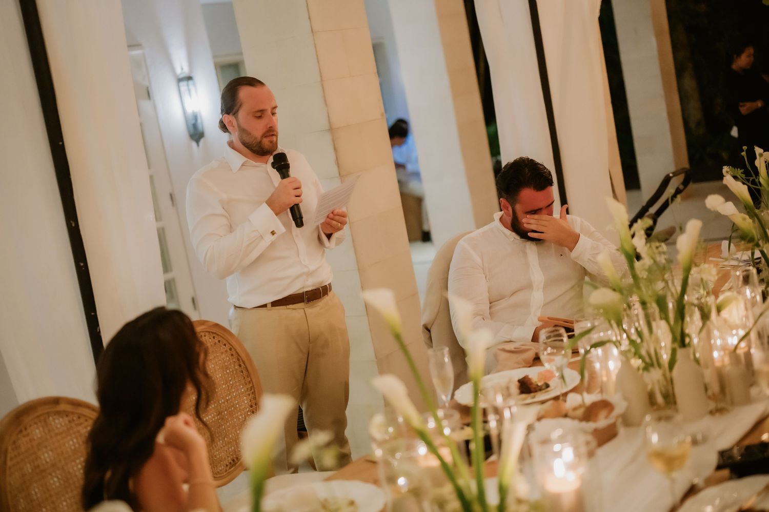 Guest delivers a heartfelt speech at candlelit wedding reception dinner table with white floral arrangements.