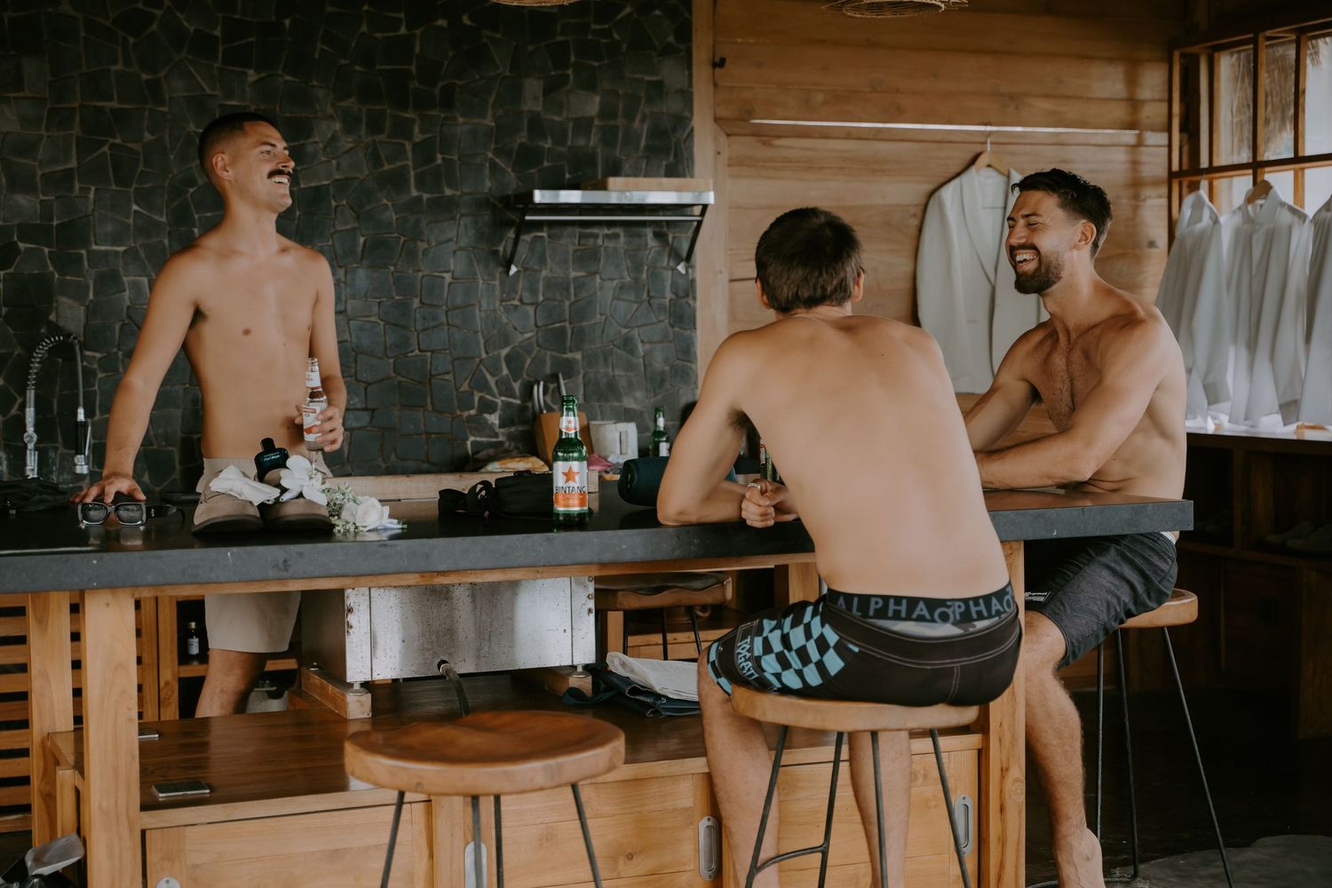 Three shirtless groomsmen laughing and celebrating at a wooden bar during pre-wedding preparations.