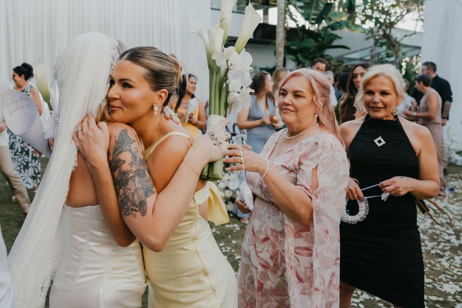 Bride in white gown with veil greeting female guests at outdoor wedding reception.