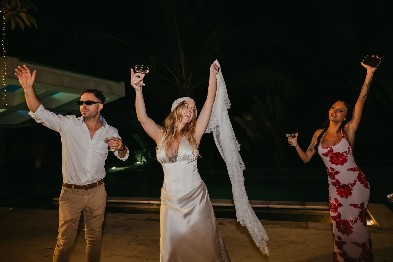 Bride in white gown and veil celebrates outdoors at night with two guests raising drinks and arms in joy.